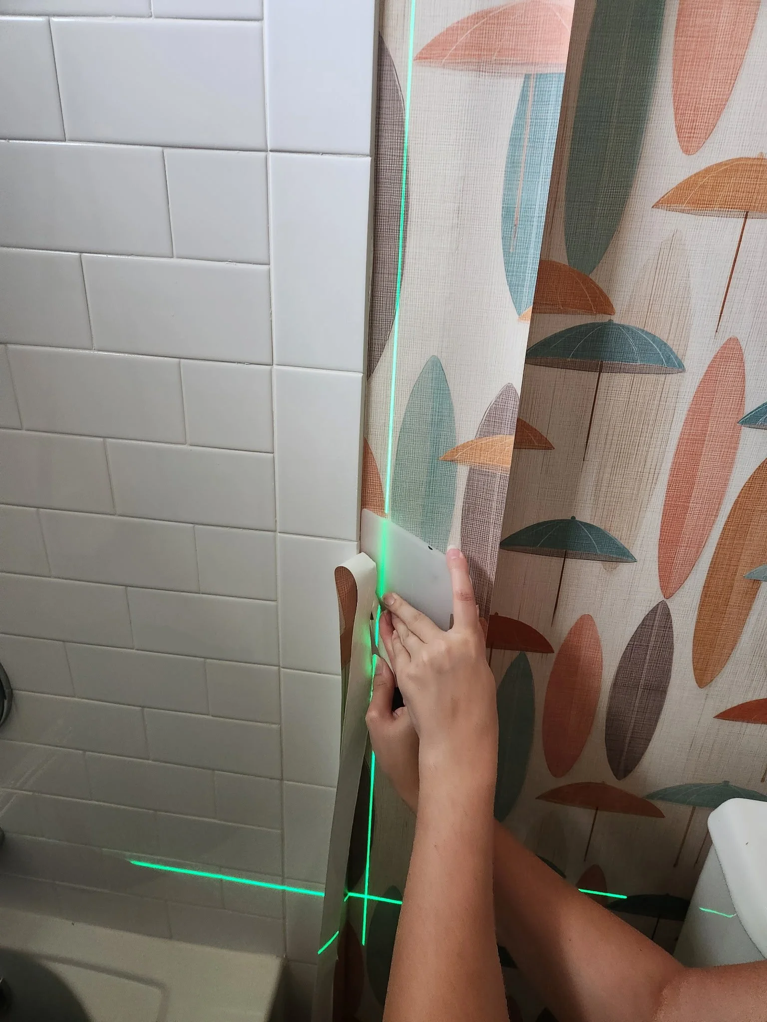 Person using a green laser level to align a panel beside bathroom wall tiles and a patterned shower curtain