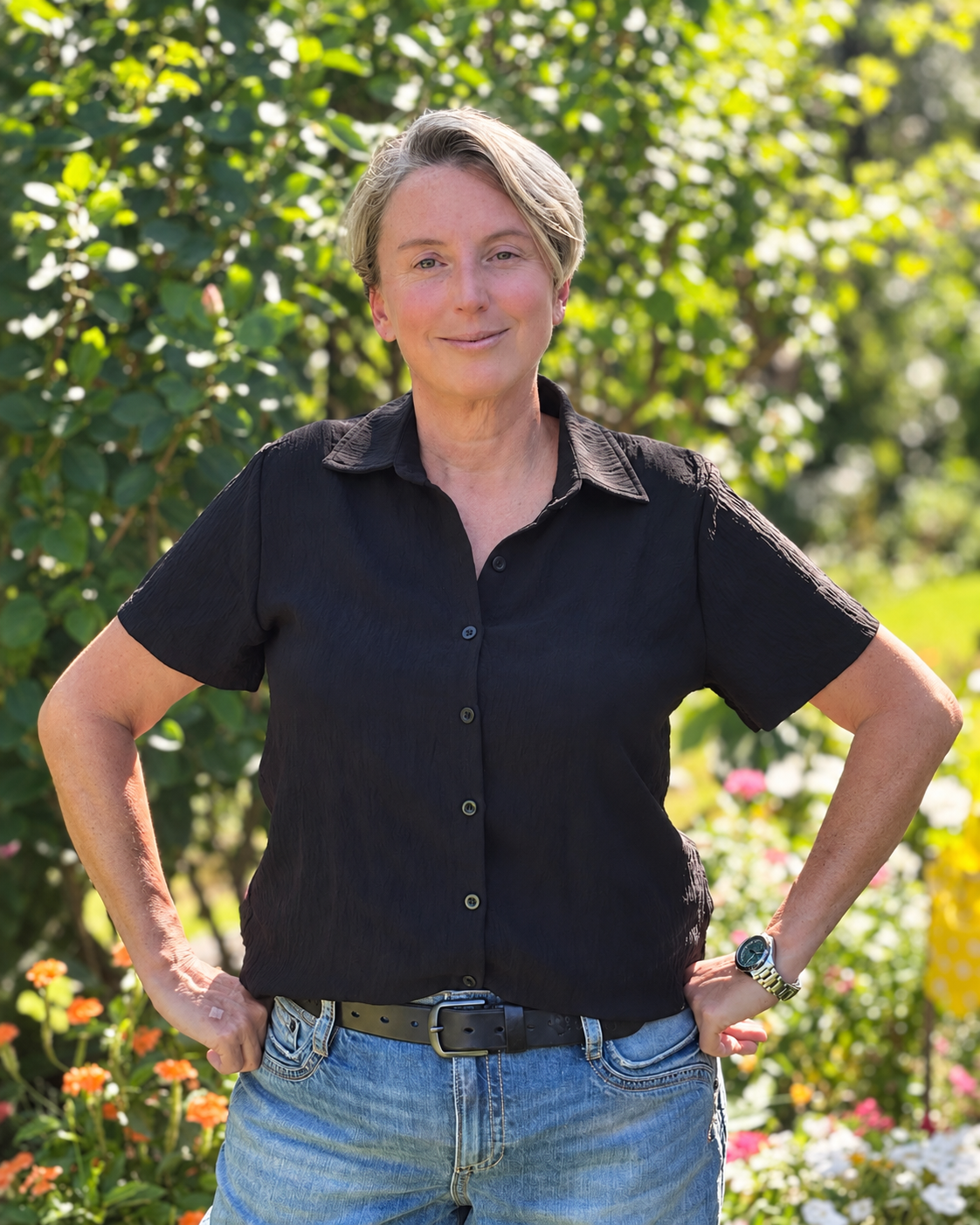 April of Redesigning Rooms smiling in a black shirt and jeans standing with hands on hips in a sunny flower garden