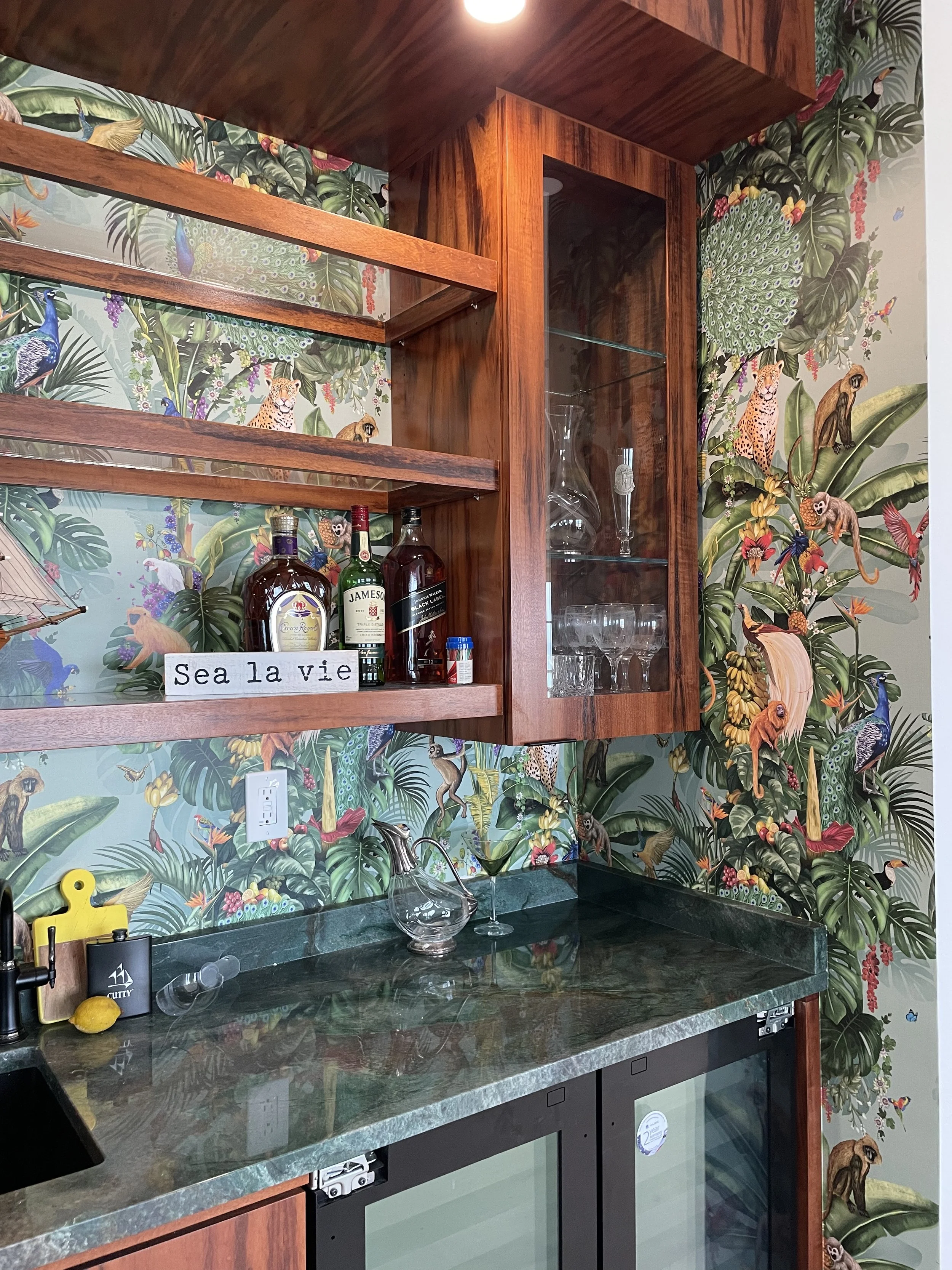 Home bar with liquor bottles, glassware, dark stone countertop, wooden shelves, and colorful tropical jungle wallpaper featuring peacocks and wild animals.