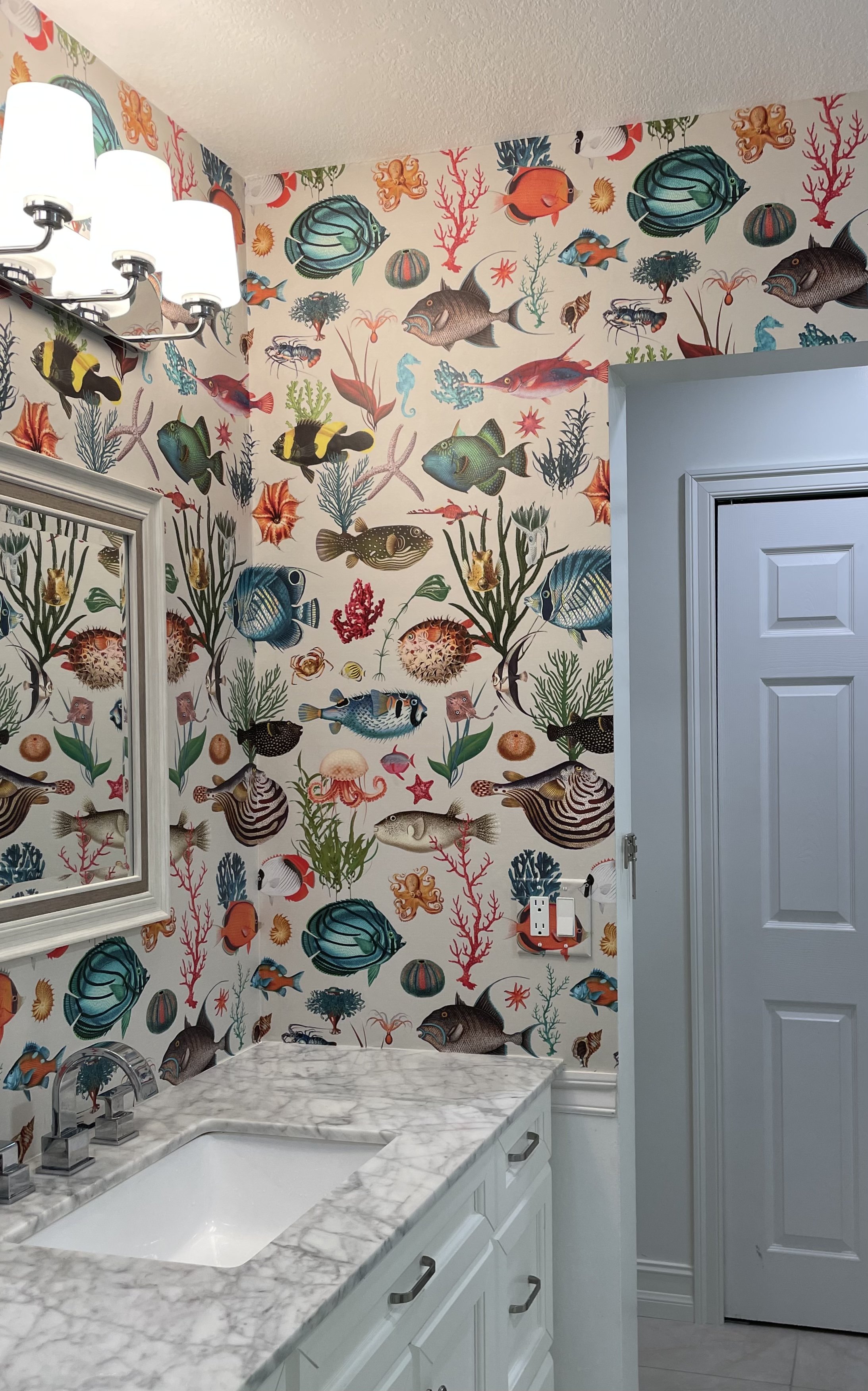 Bright bathroom with white marble vanity, bathtub, chandelier, and colorful vintage ocean-themed wallpaper featuring fish, coral, octopuses, and sea life.