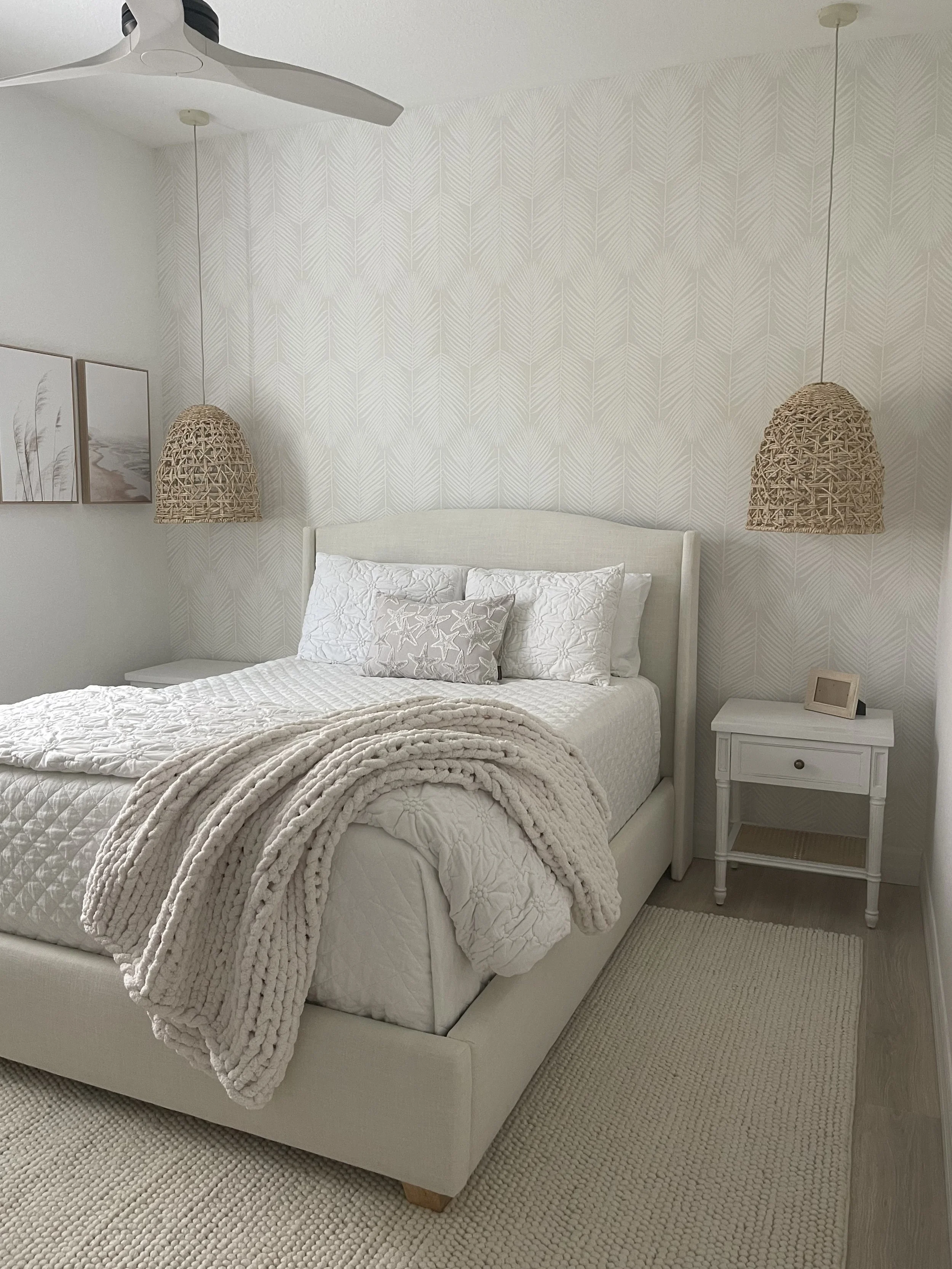 Bright white bedroom with upholstered bed, textured bedding, woven pendant lights, patterned wallpaper, ceiling fan, framed artwork, and cream area rugs.