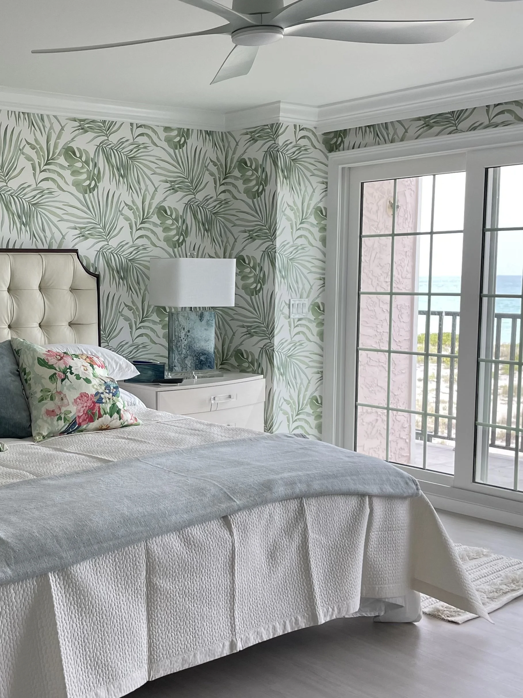 Bright coastal bedroom with tropical leaf wallpaper, white bed, blue lamp, ceiling fan, and French doors opening to a balcony.