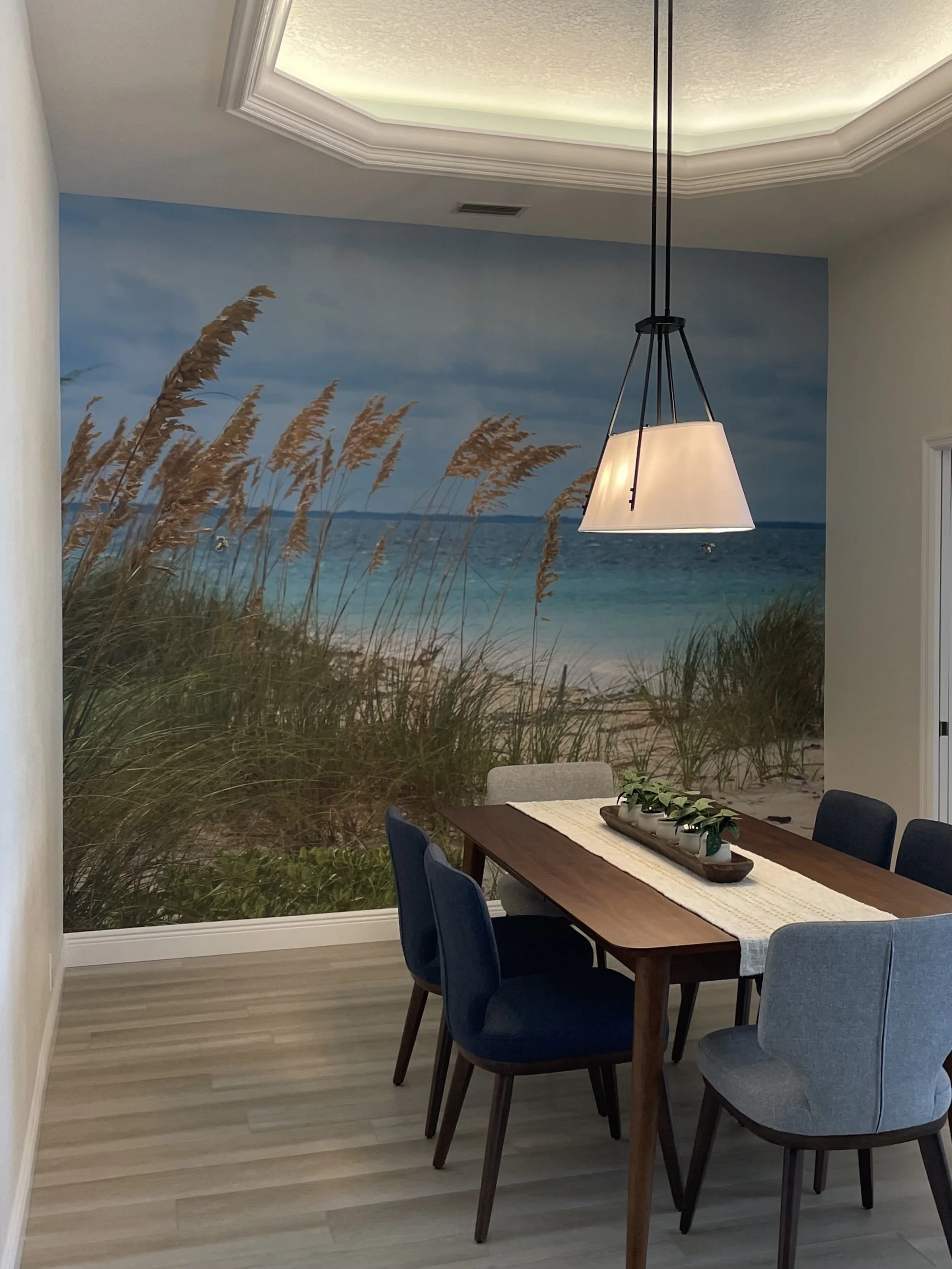 Coastal dining room with blue upholstered chairs, wooden table, pendant light, and beach mural featuring dune grass and ocean