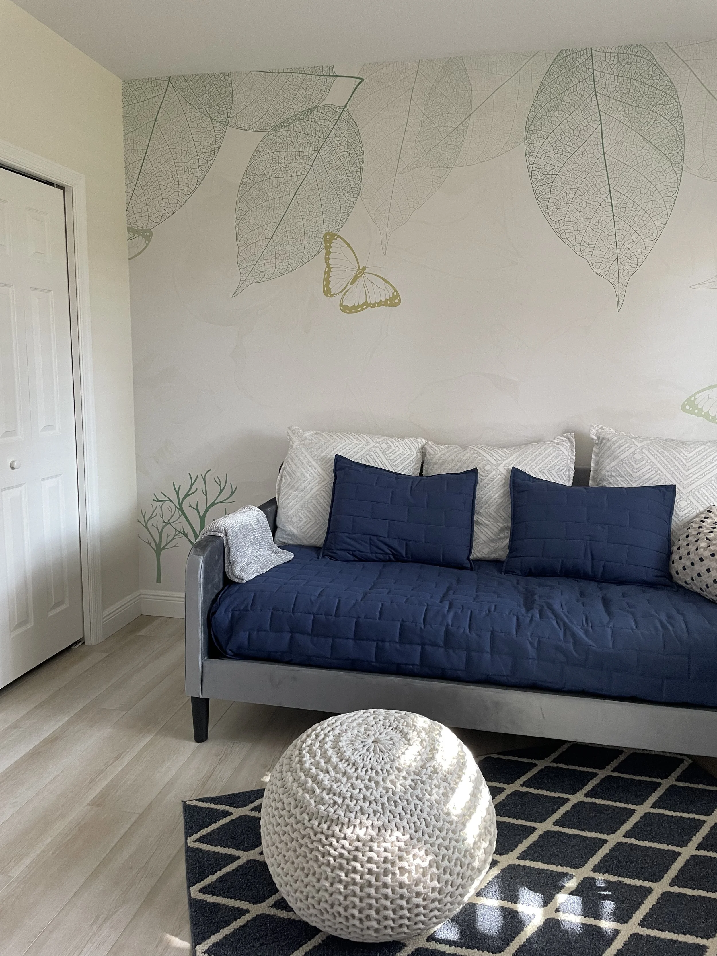 Bright living room with a gray daybed, navy and white pillows, oversized leaf and butterfly wall art, a white knitted pouf, and black geometric rug.