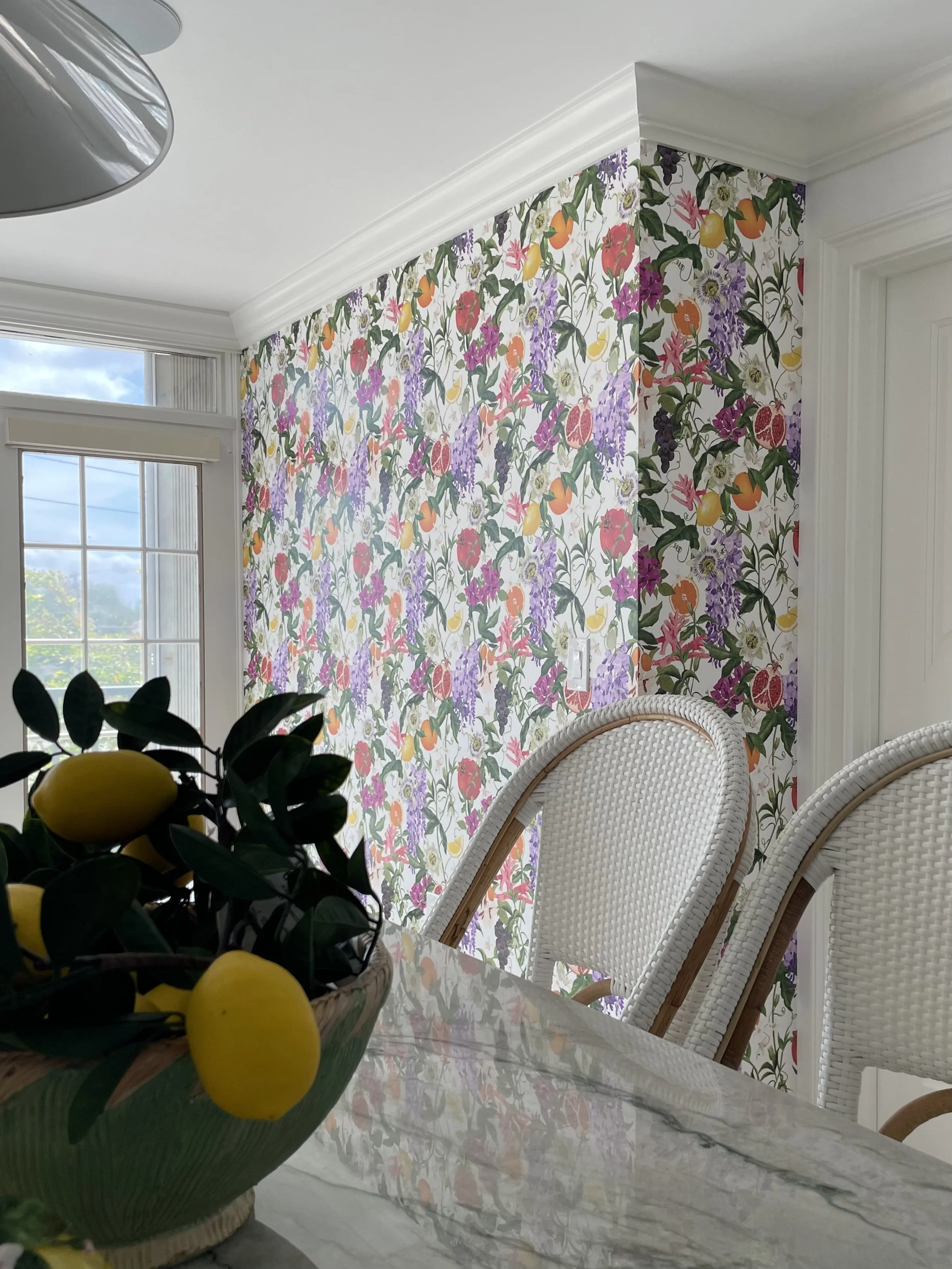 Bright dining area featuring colorful floral and fruit wallpaper, white wicker chairs, and a marble table.