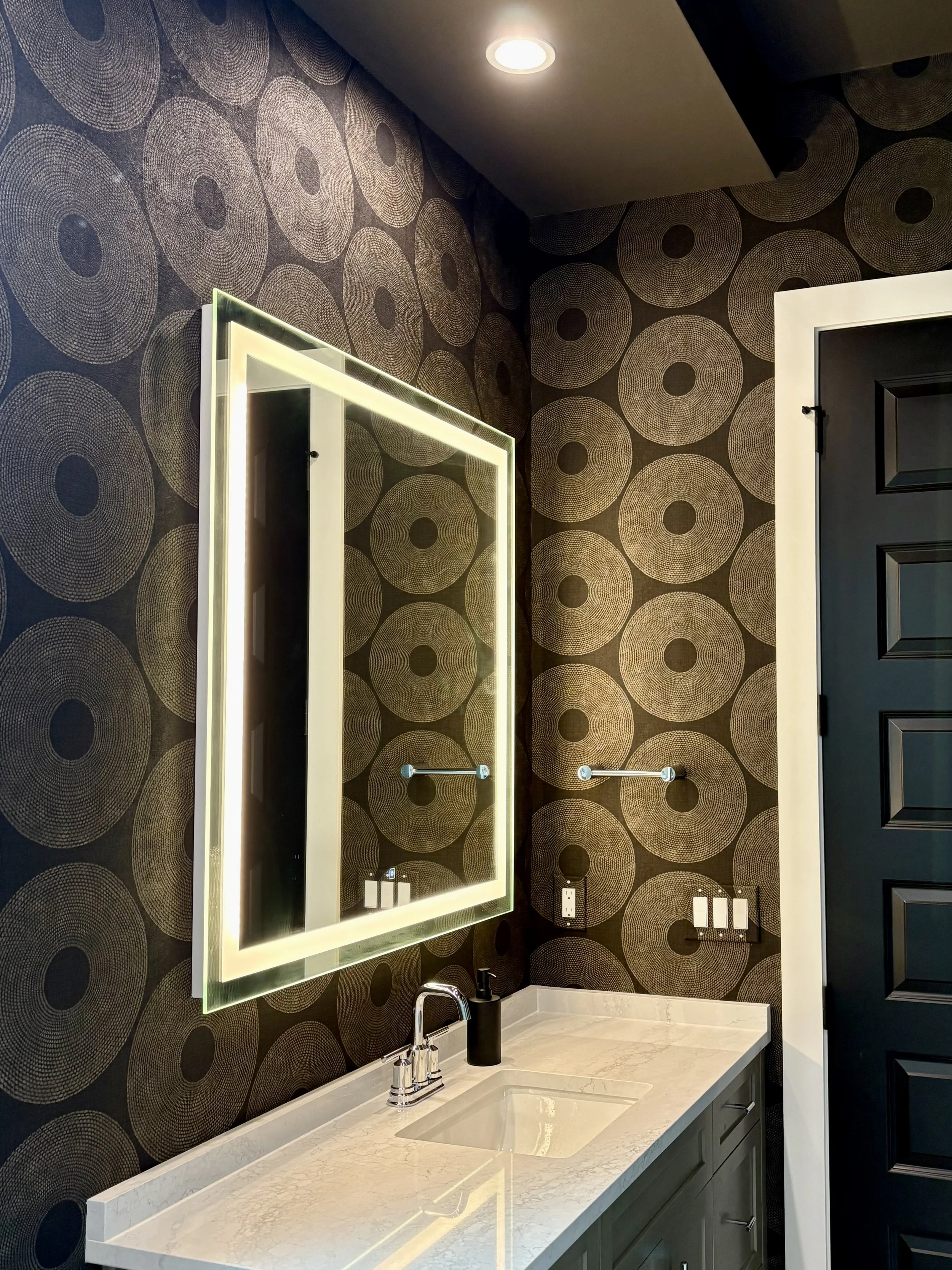 Modern powder room with dark circular-pattern wallpaper, illuminated mirror, marble vanity, chrome faucet, and black paneled door