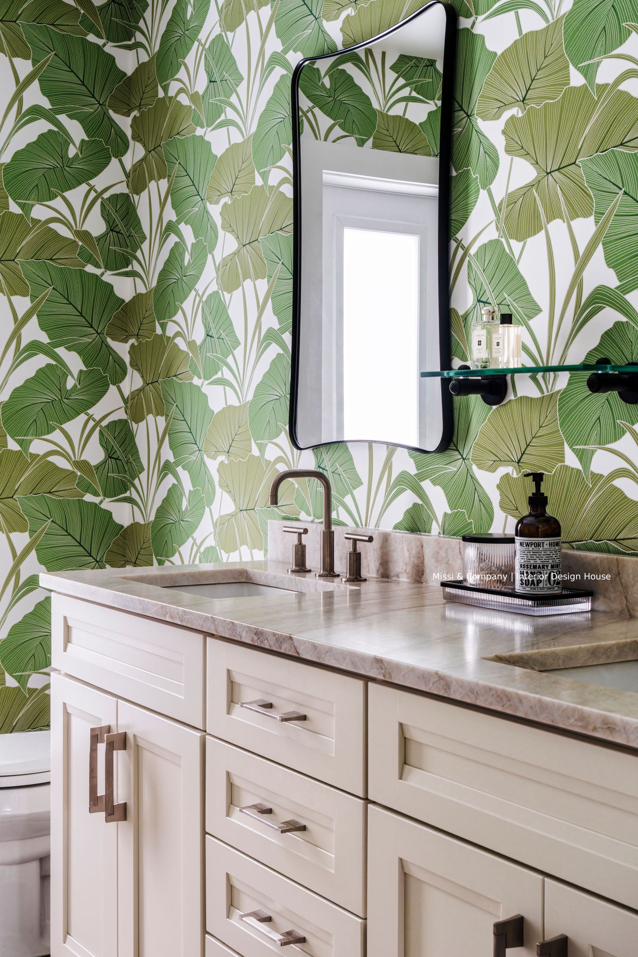 Elegant bathroom vanity with cream cabinets, marble countertop, black-framed mirror, glass shelf, and green tropical leaf wallpaper