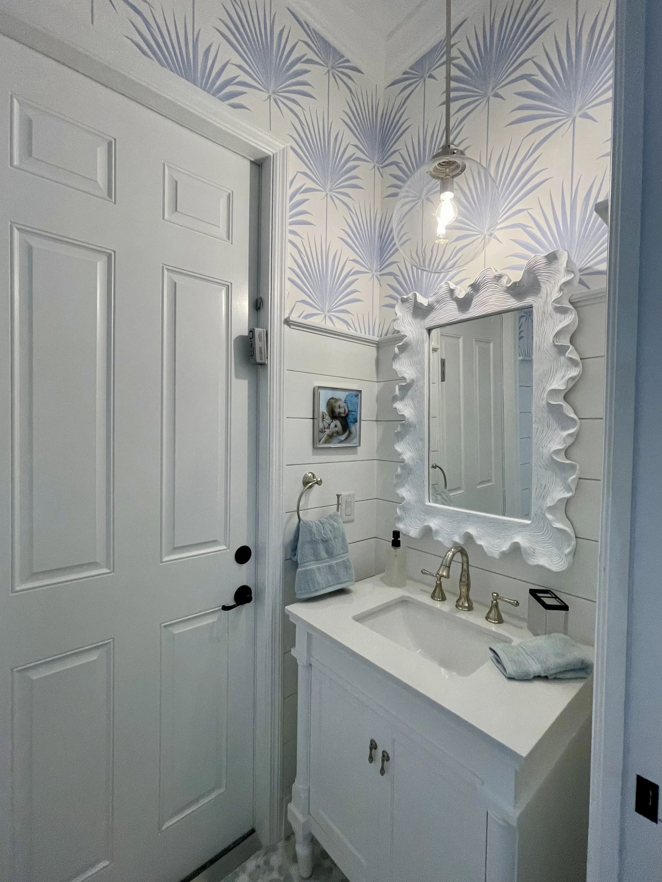 Bathroom with blue palm-leaf wallpaper, decorative shell-framed mirror, brass fixtures, and white vanity.