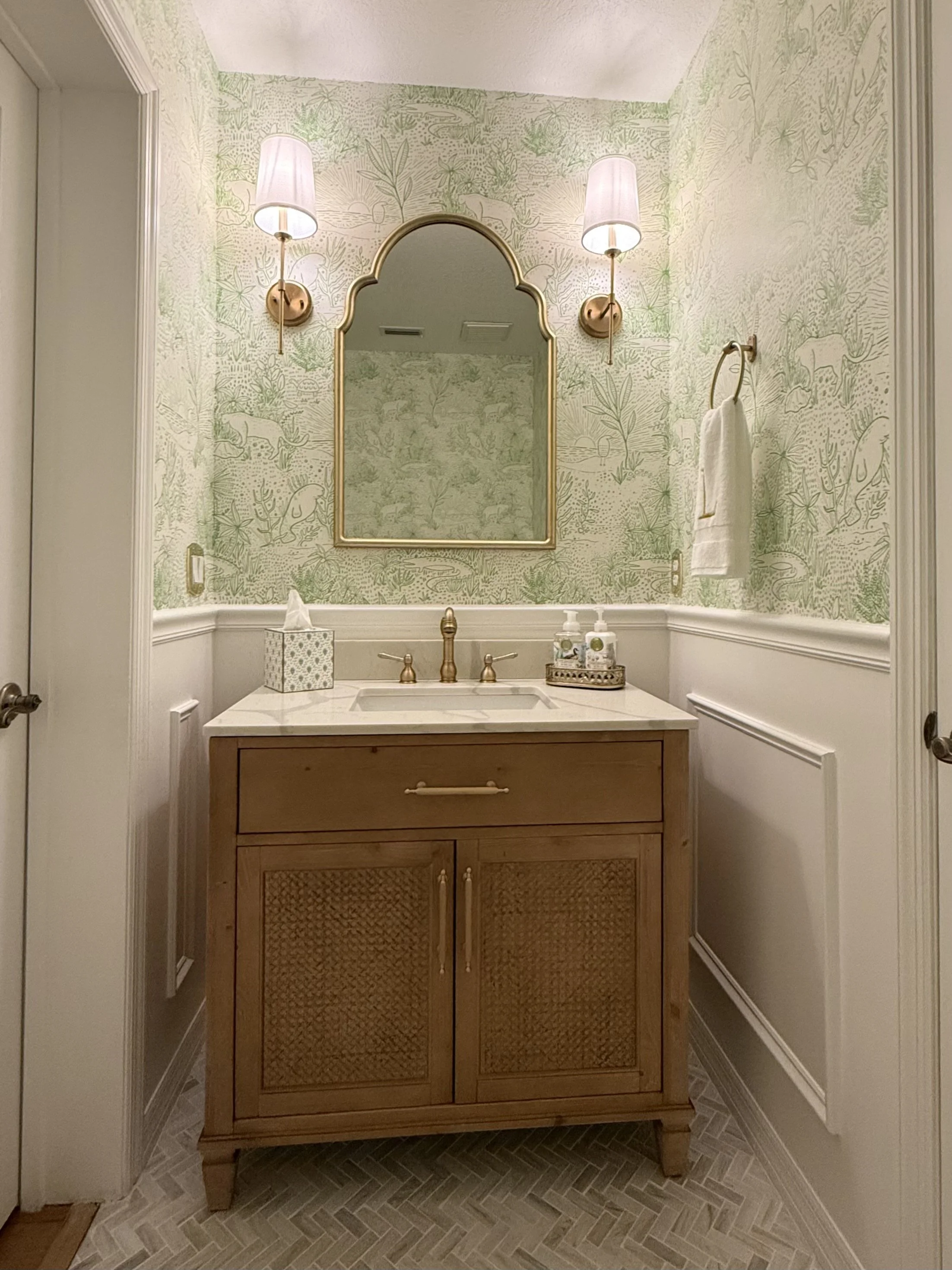 Elegant powder bathroom with green jungle-animal wallpaper, arched gold-framed mirror, brass sconces, wood vanity, and herringbone tile floor