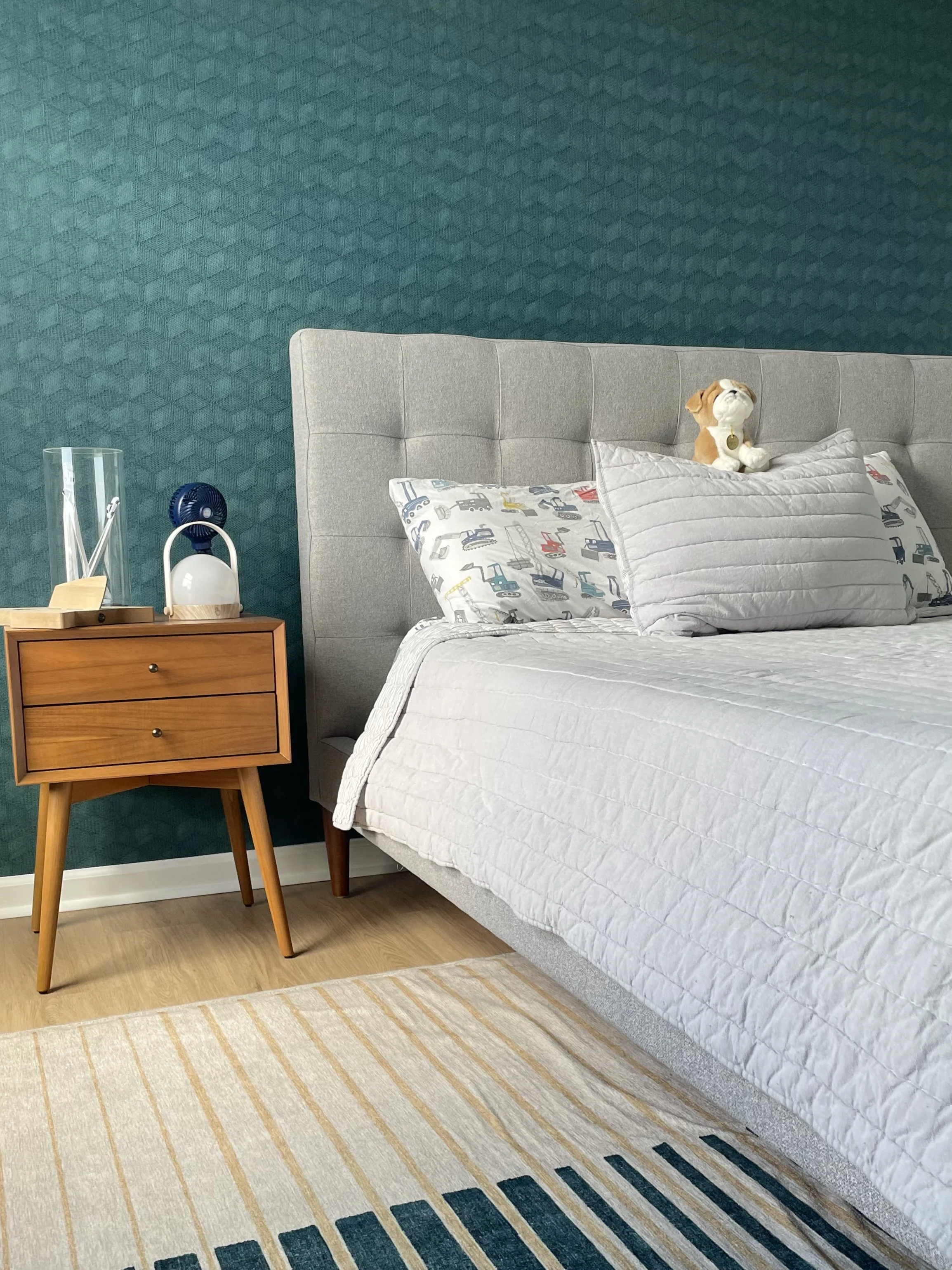 Cozy child’s bedroom with a gray bed, construction-vehicle bedding, bulldog stuffed animal, teal geometric wallpaper, and wooden nightstand