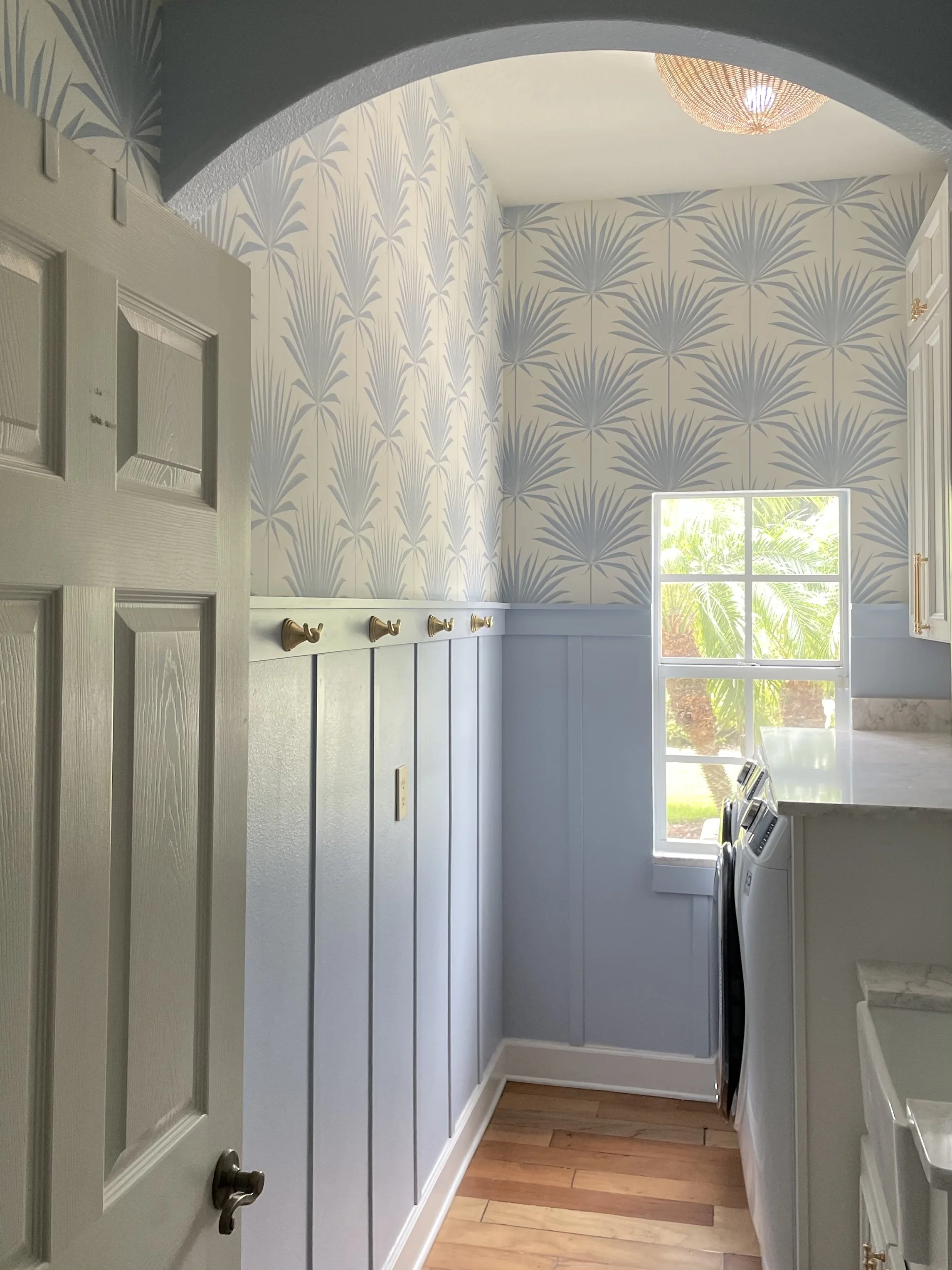 Bright laundry room with blue palm-leaf wallpaper, beadboard paneling, brass hooks, pendant light, window, washer, and dryer.