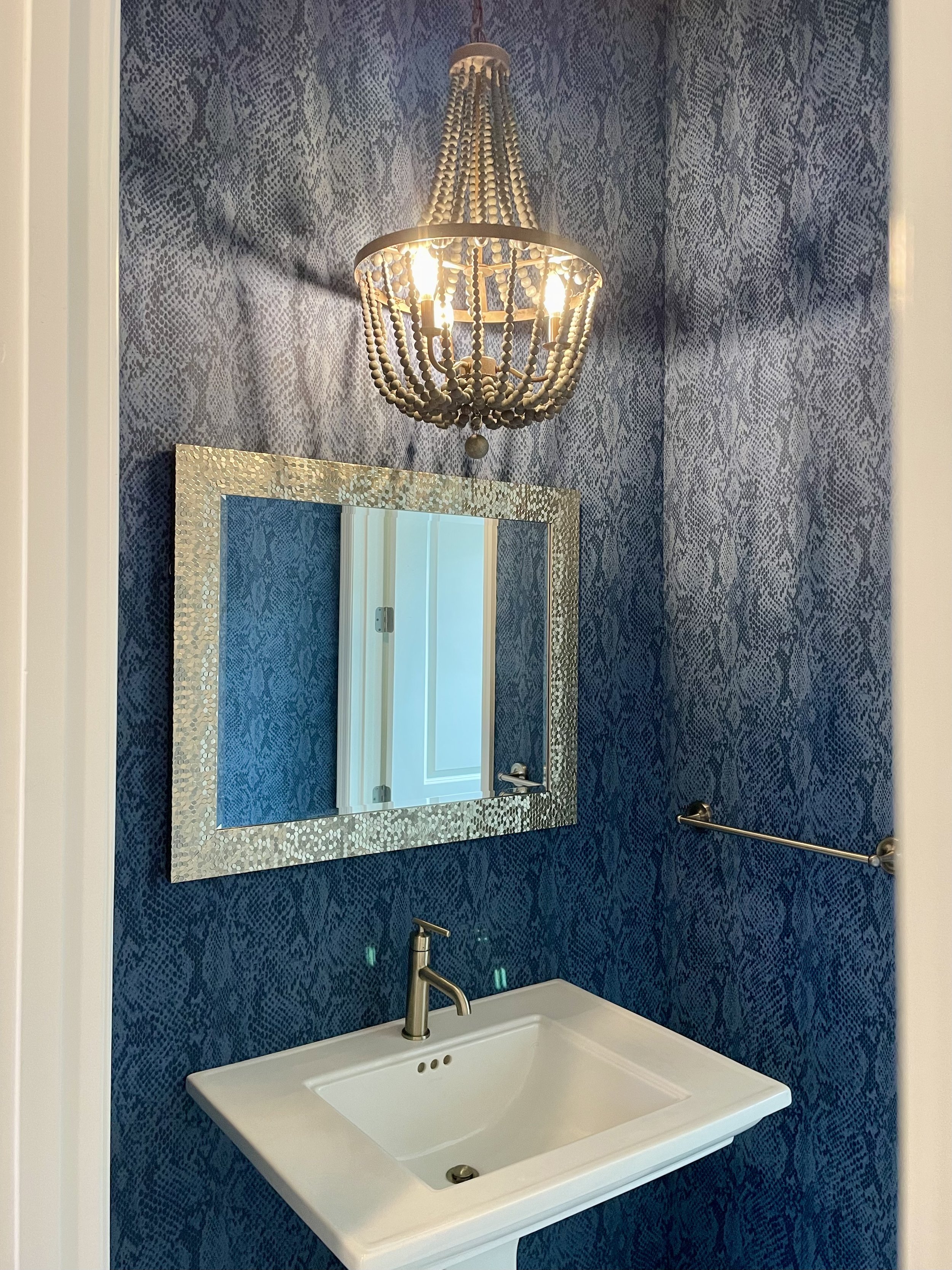 Elegant bathroom vanity with white pedestal sink, silver textured mirror, blue patterned wallpaper, towel bar, and illuminated beaded chandelier
