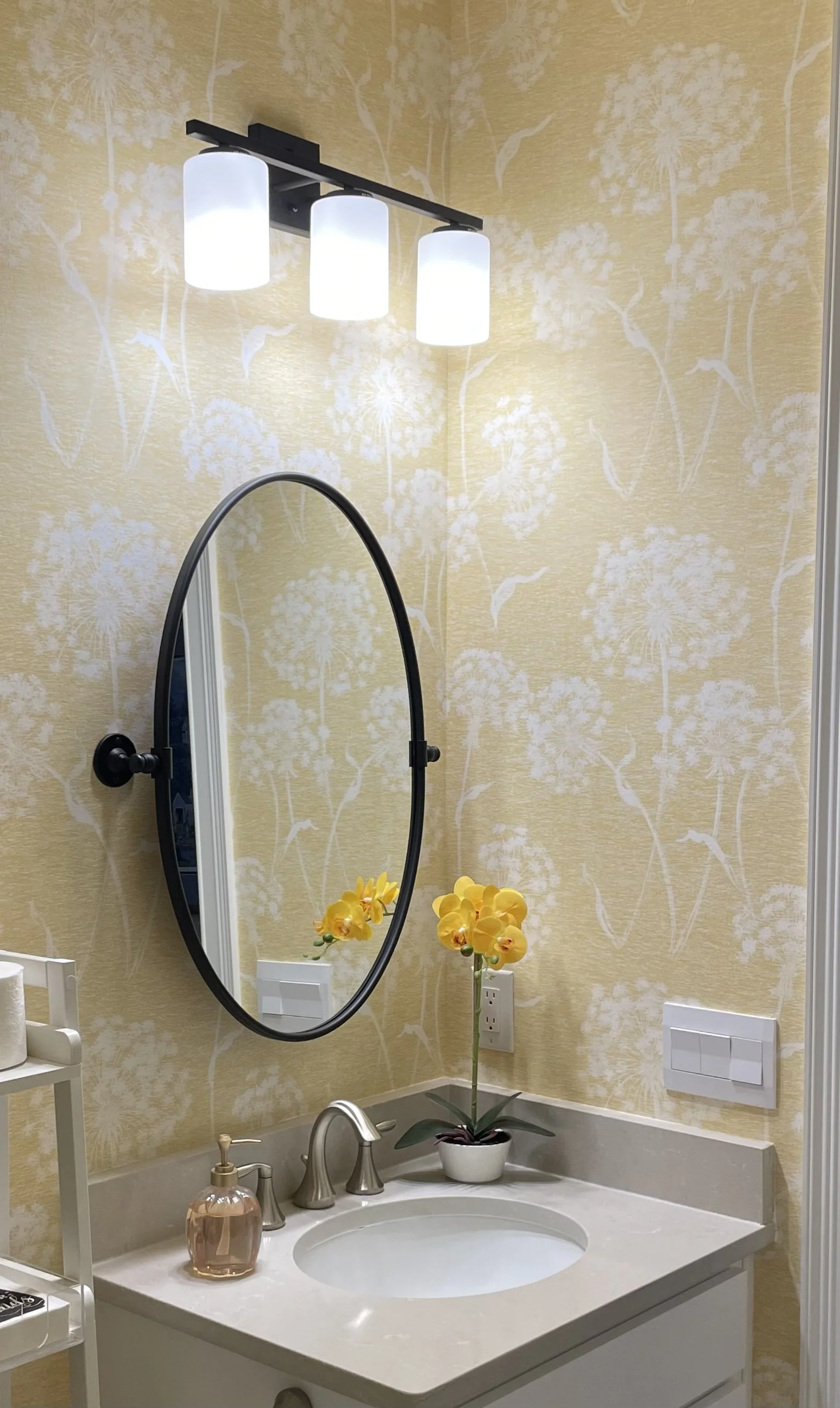 Modern bathroom vanity with oval mirror, three-light fixture, white sink, silver faucet, and yellow orchid
