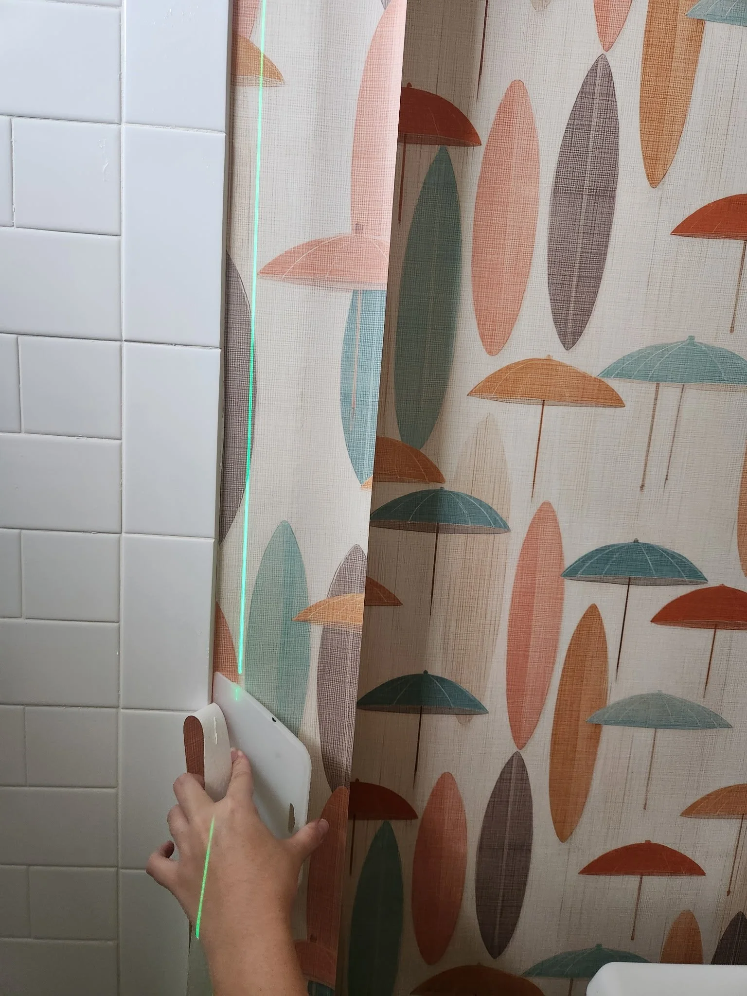 Installer using a laser level to align colorful botanical wallpaper beside white bathroom tile.