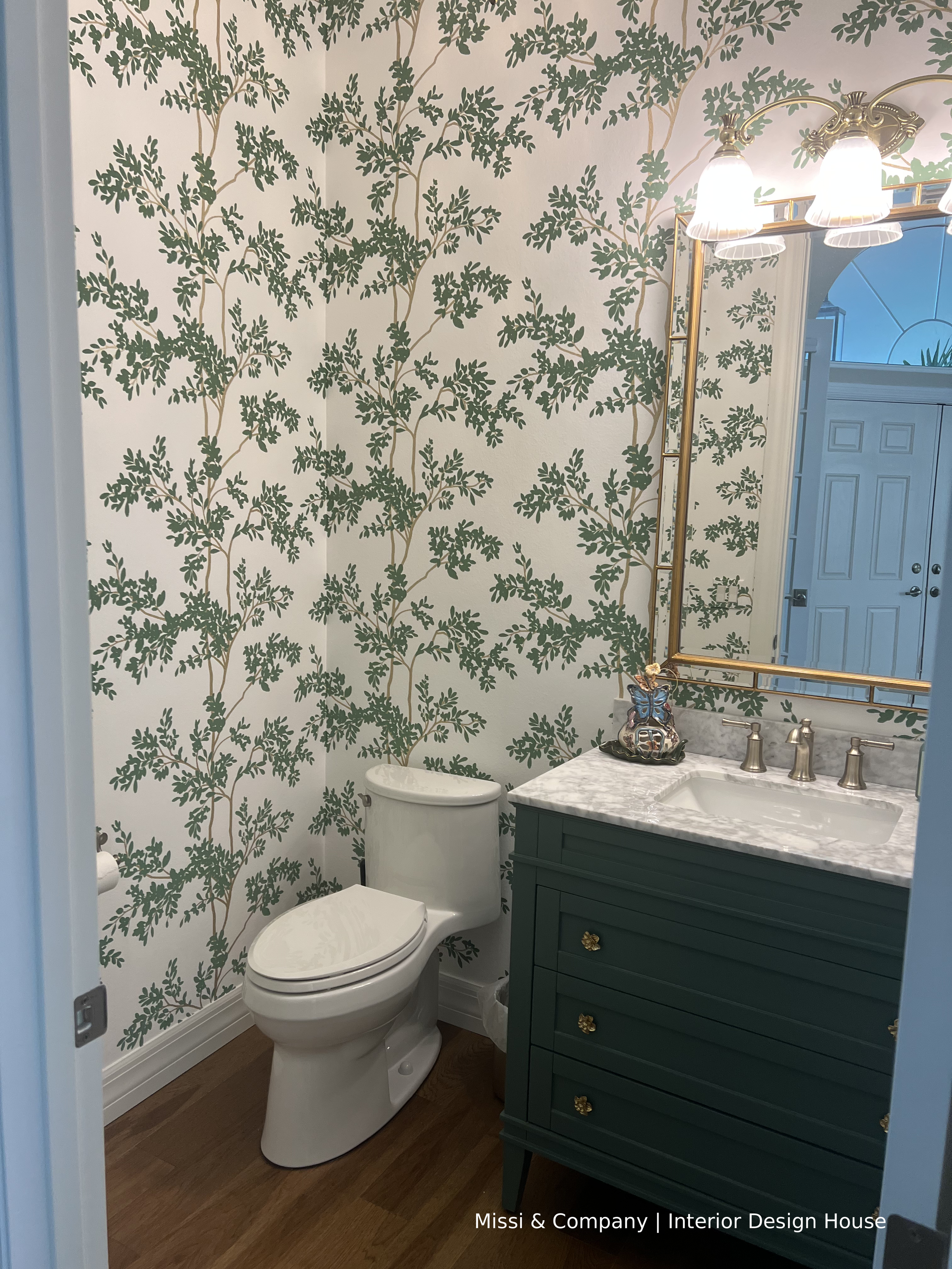 Powder room featuring green botanical wallpaper, dark green vanity, marble countertop, and brass-framed mirror.