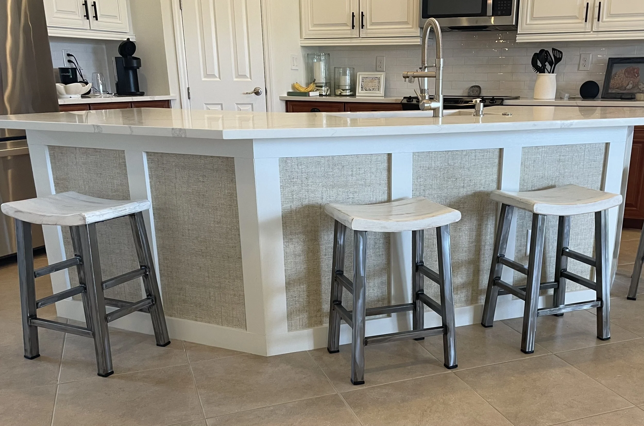 Bright kitchen with a white island, four upholstered bar stools, stainless steel appliances, and a farmhouse sink.