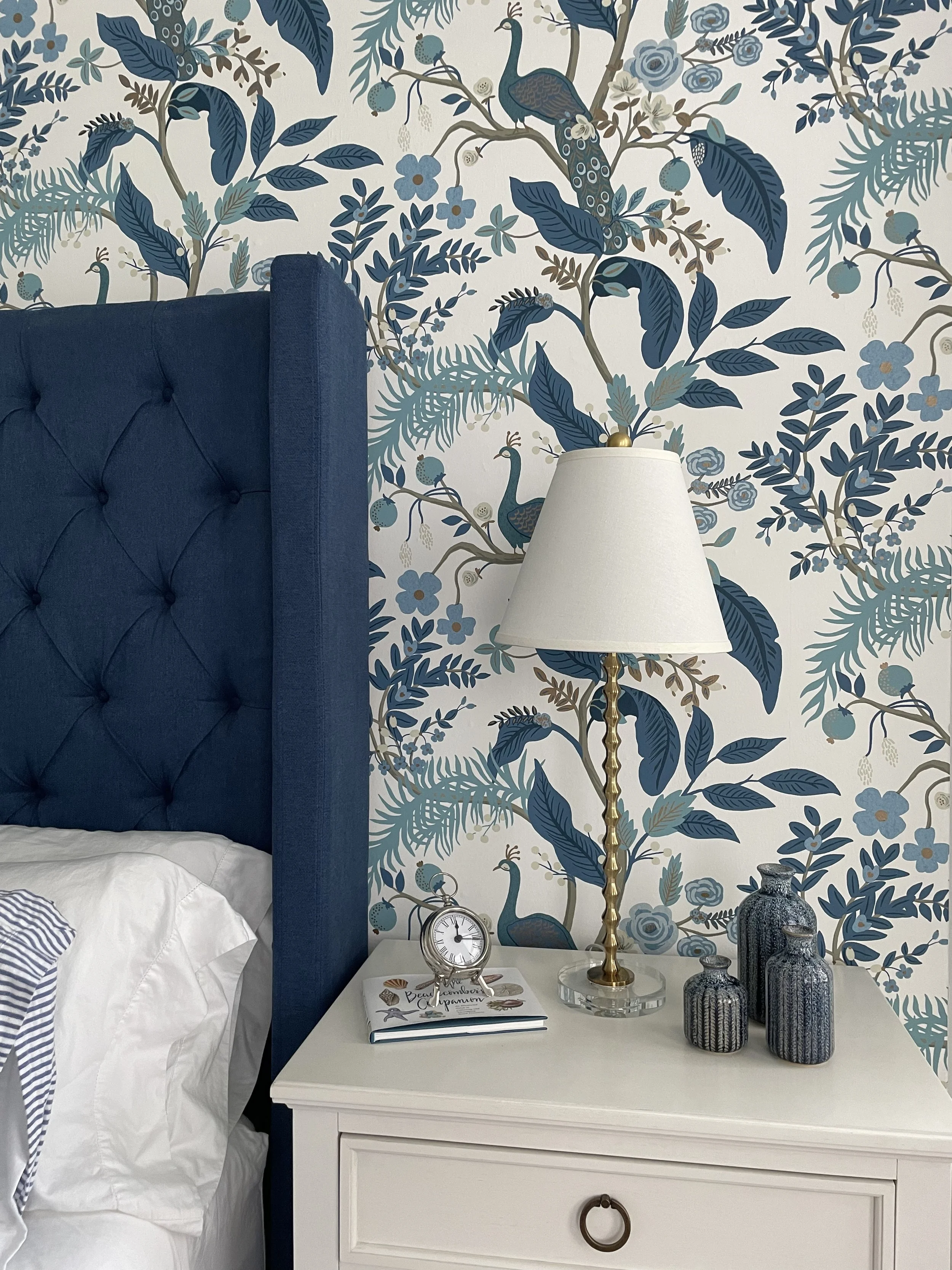 Elegant bedroom with blue tufted headboard, peacock and floral wallpaper, white nightstand, brass lamp, clock, book, and decorative vases.