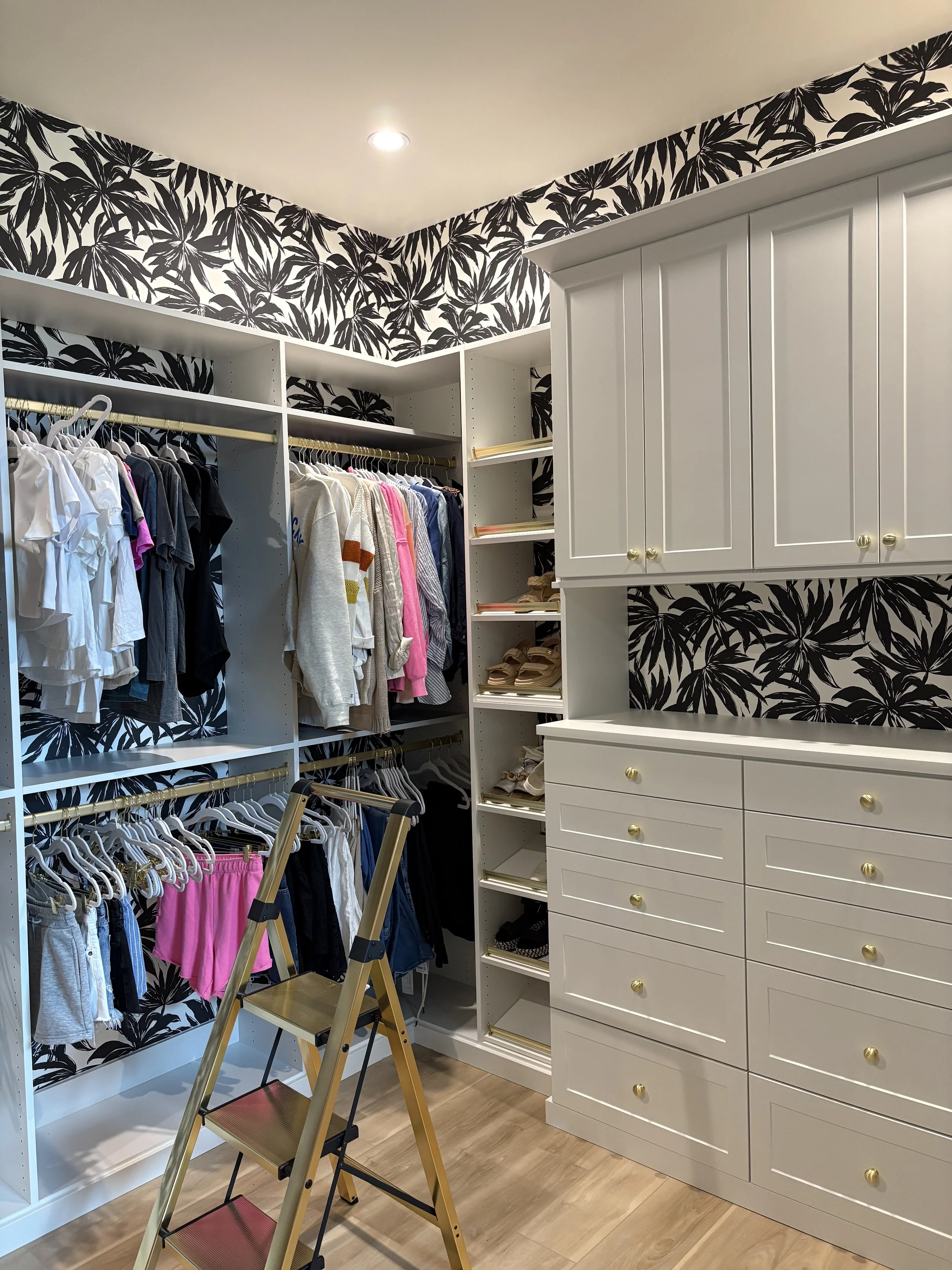 Custom walk-in closet with black-and-white tropical leaf wallpaper, white shelves and drawers, hanging clothes, shoe storage, and a yellow step ladder.