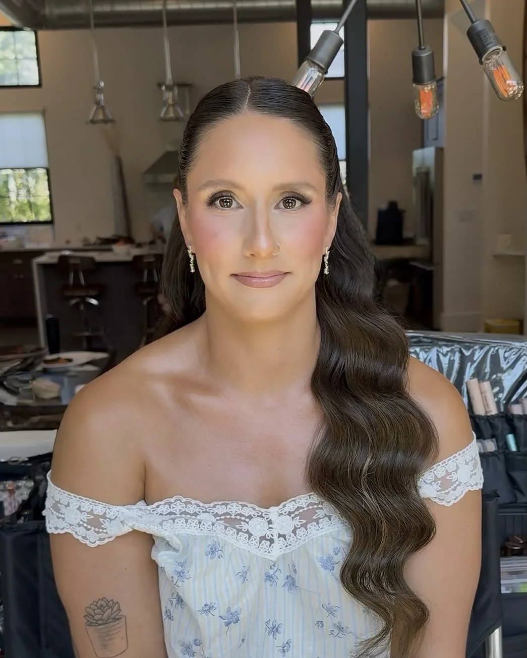 Woman with long wavy hair, wearing a white off-shoulder dress with lace and floral patterns, and earrings, in a well-lit, modern indoor space.