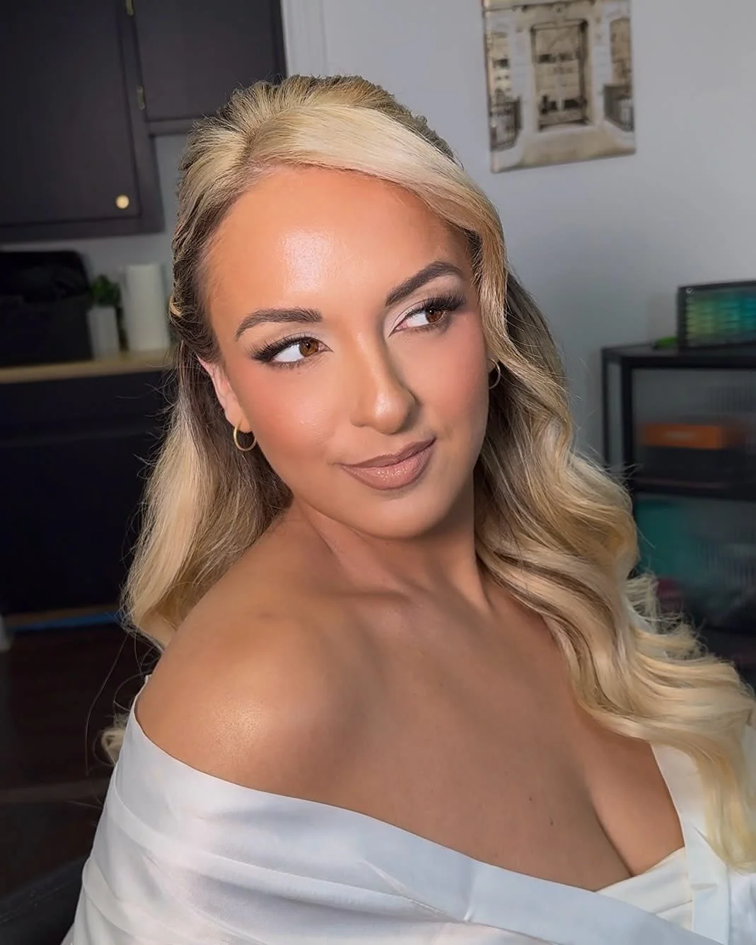 Close-up of a woman with long blond wavy hair, wearing makeup and a white off-the-shoulder top, in a room with dark cabinets and a framed picture on the wall.