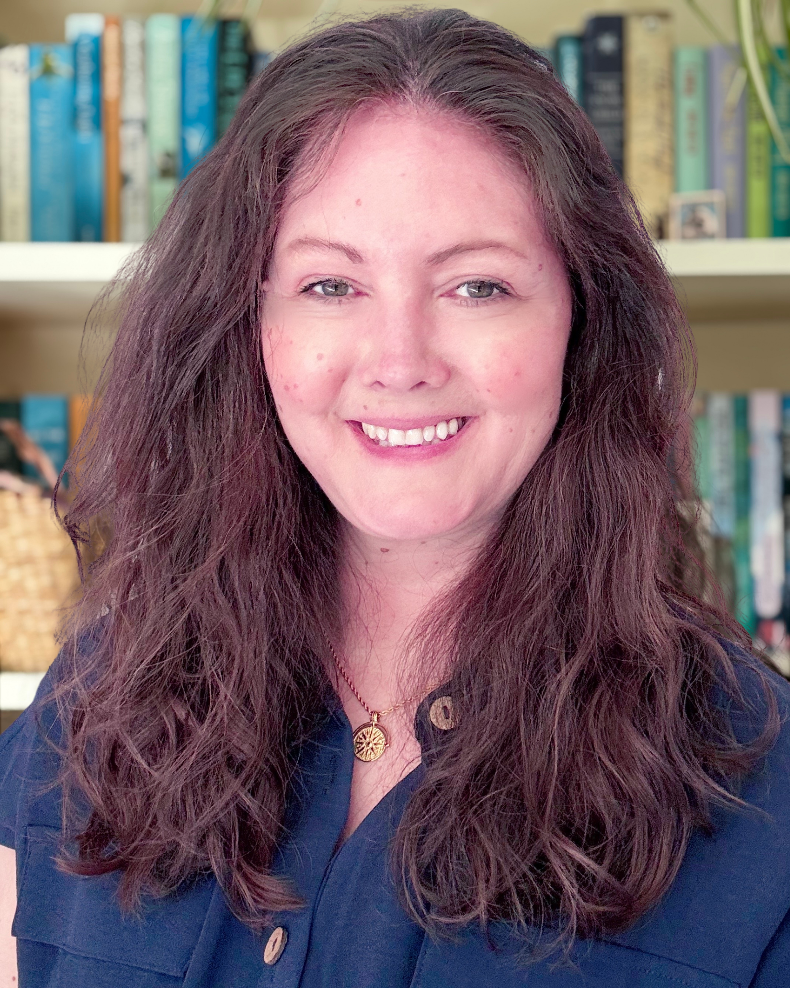 Smiling woman with long dark hair wearing a navy blouse and pendant necklace in front of a bookshelf