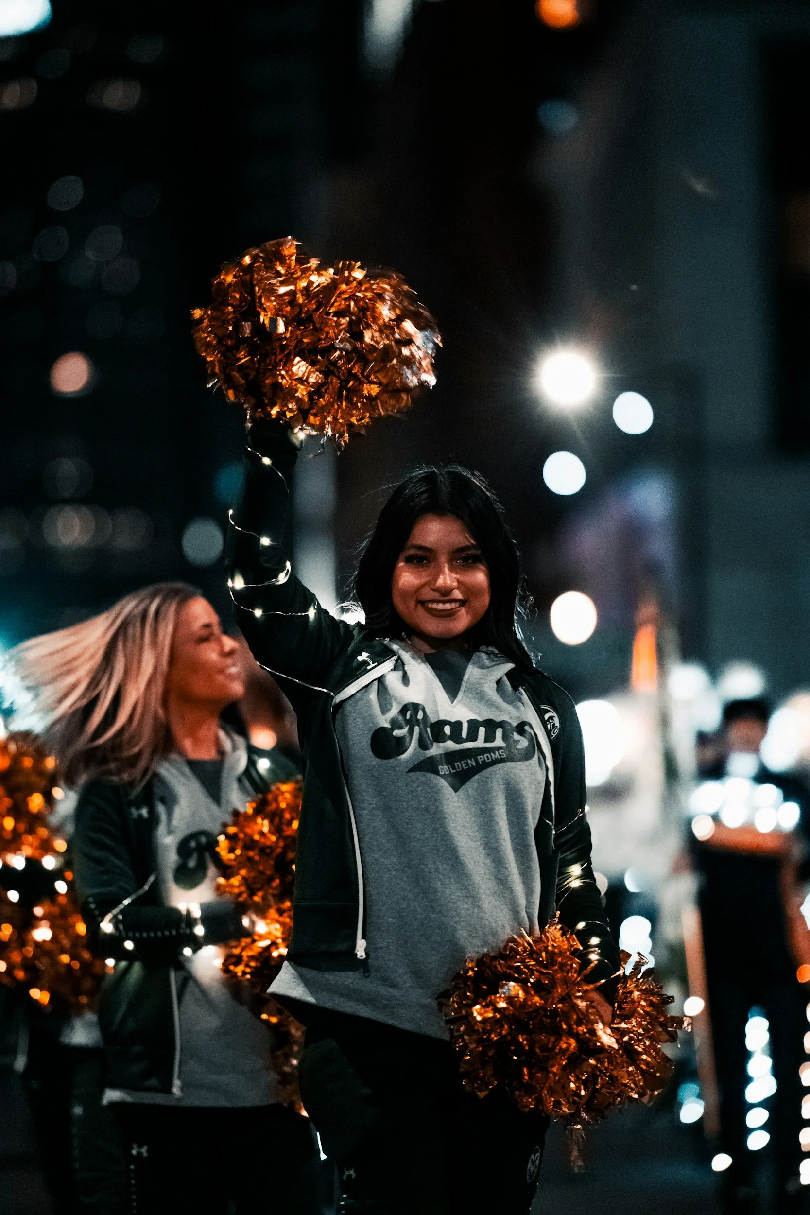 Group of cheerleaders in uniform holding orange pom-poms, smiling, in an urban nighttime setting with city lights and traffic in the background.