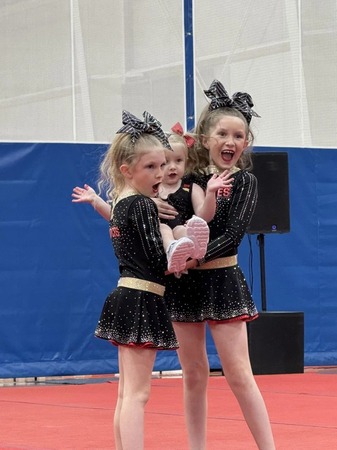 Three young girls in black dance costumes with sparkly embellishments, bows in their hair, standing on a red mat inside a gymnasium, one girl holding another, all showing excited expressions.