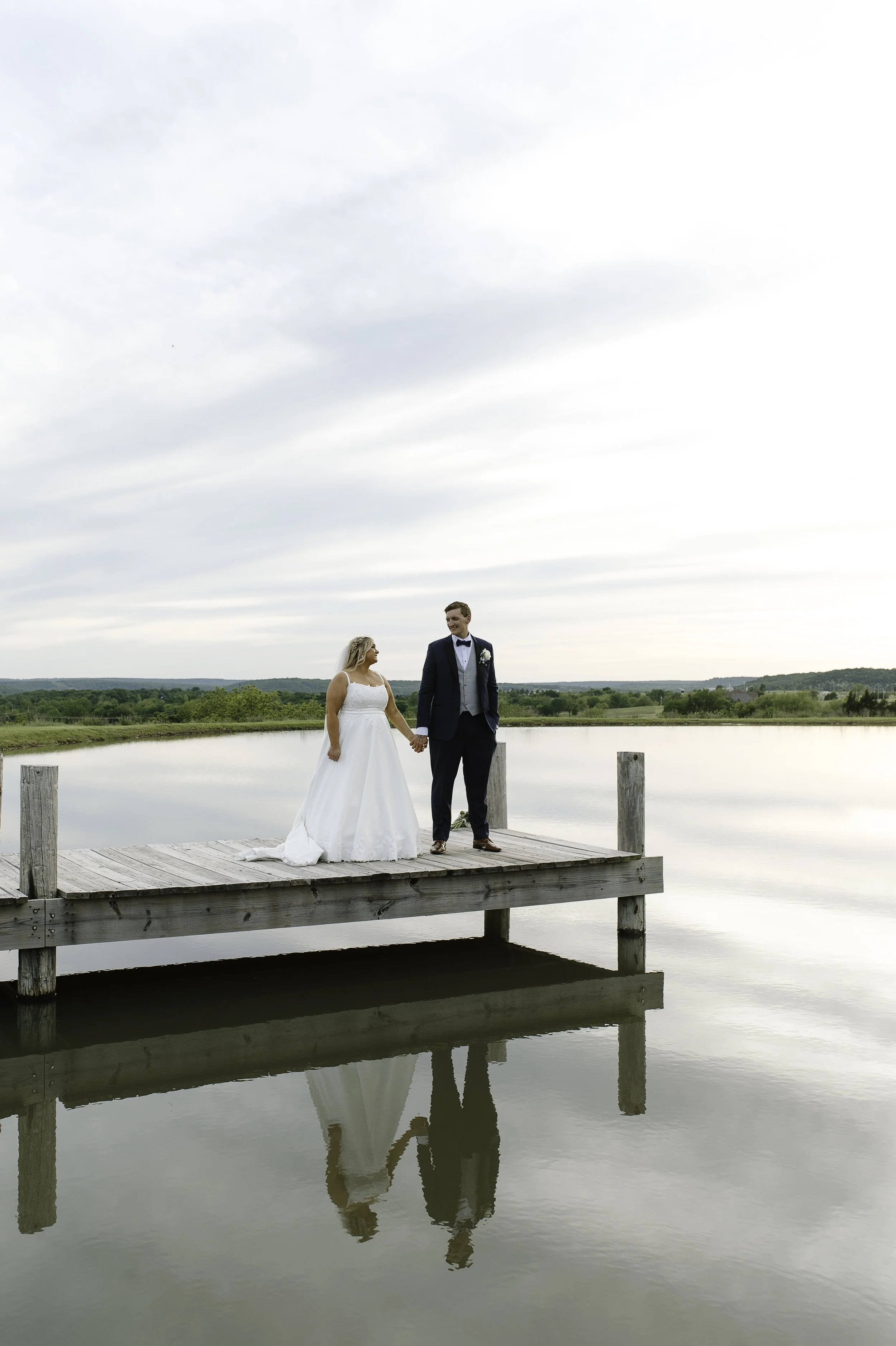 A bride and groom walking hand in hand on a wooden dock over a calm lake during sunset, with trees and green hills in the background.