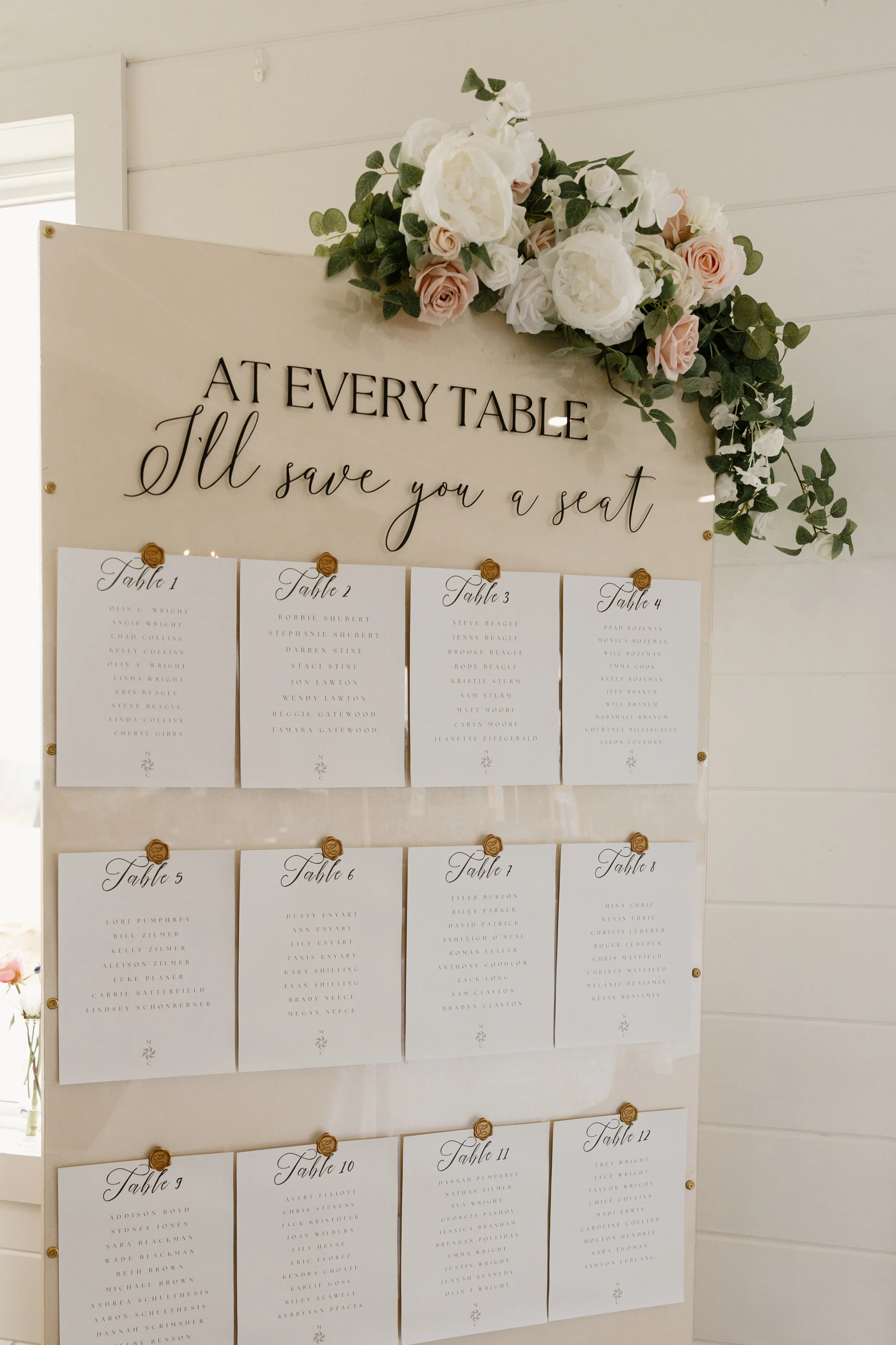 A wedding seating chart displayed on a wall, with a floral arrangement of white and blush roses and greenery at the top. The chart has a heading that says 'AT EVERY TABLE I'll save you a seat,' with numbered sheets listing guests' names organized by 