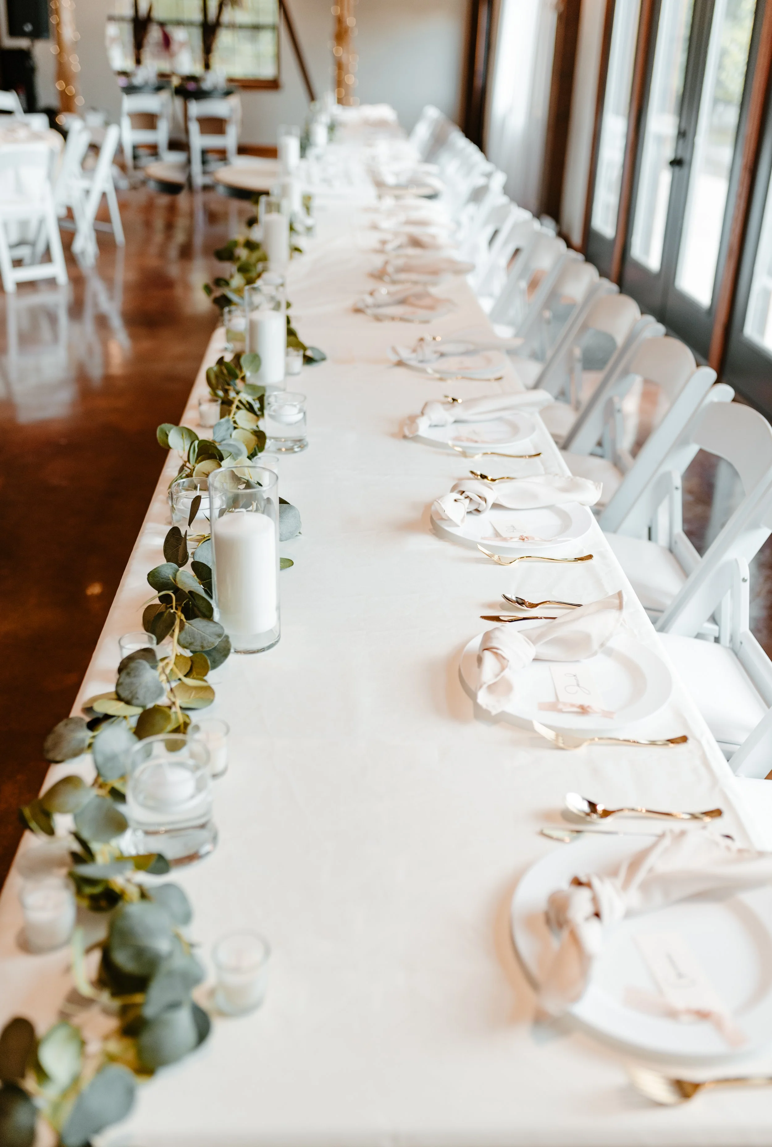 Long dining table set for an event with white tablecloth, gold cutlery, white napkins, white plates, and water glasses, decorated with candles and eucalyptus garland. Sunlight visible through glass doors.