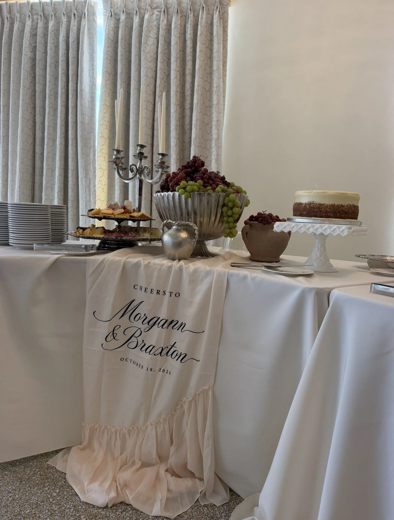 A decorated table at a wedding reception featuring a white tablecloth with a beige ruffled cloth displaying the names 'Morgan & Braxton' and the date October 18, 2026. The table holds a silver candelabrum, a large bowl of grapes, a small pot of grapes, a cake on a white pedestal, and multiple plates stacked on the side. The background has light-colored curtains.