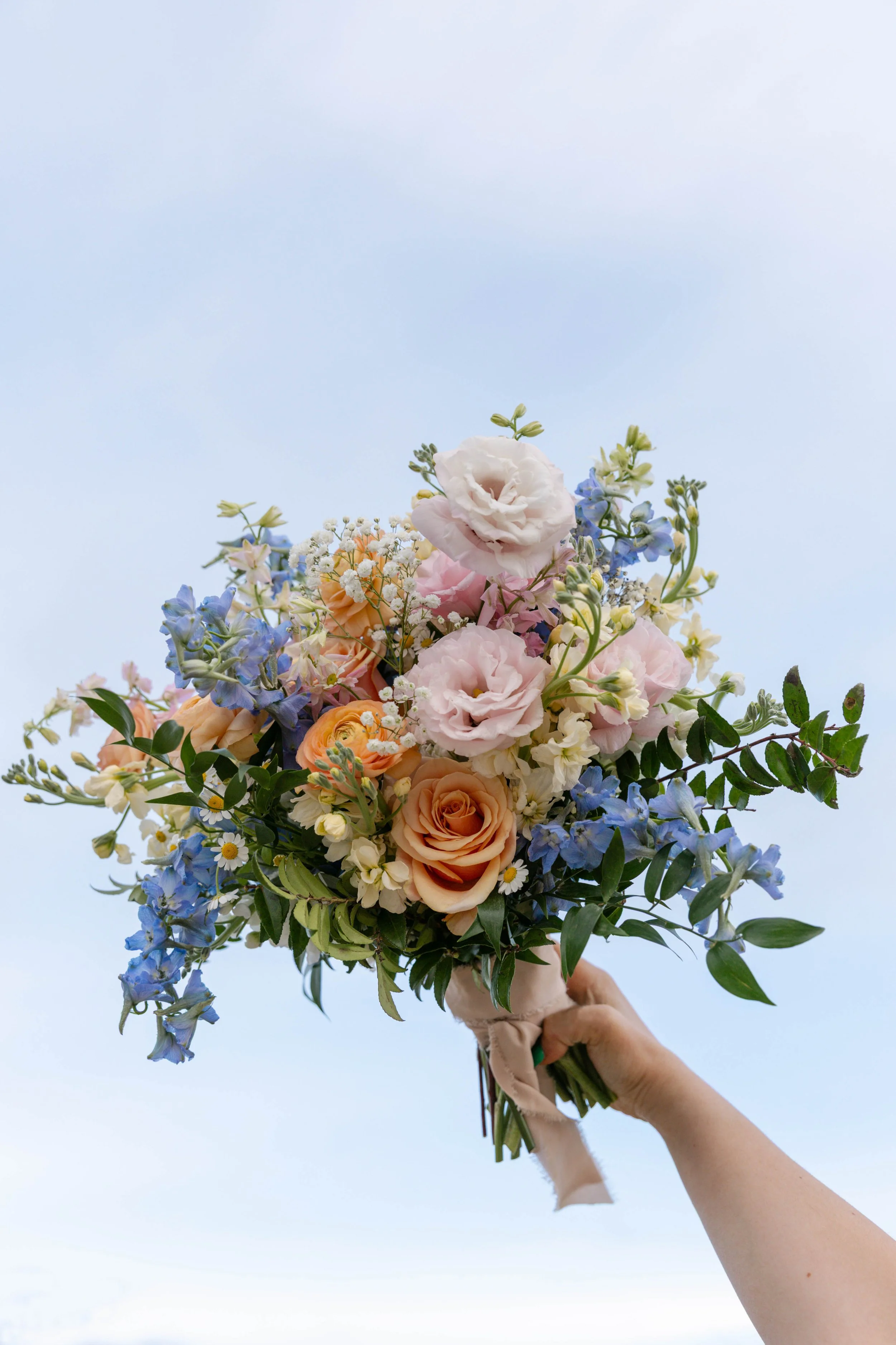 Hand holding a bouquet of mixed flowers against a clear sky, including roses, lisianthus, and daisies.