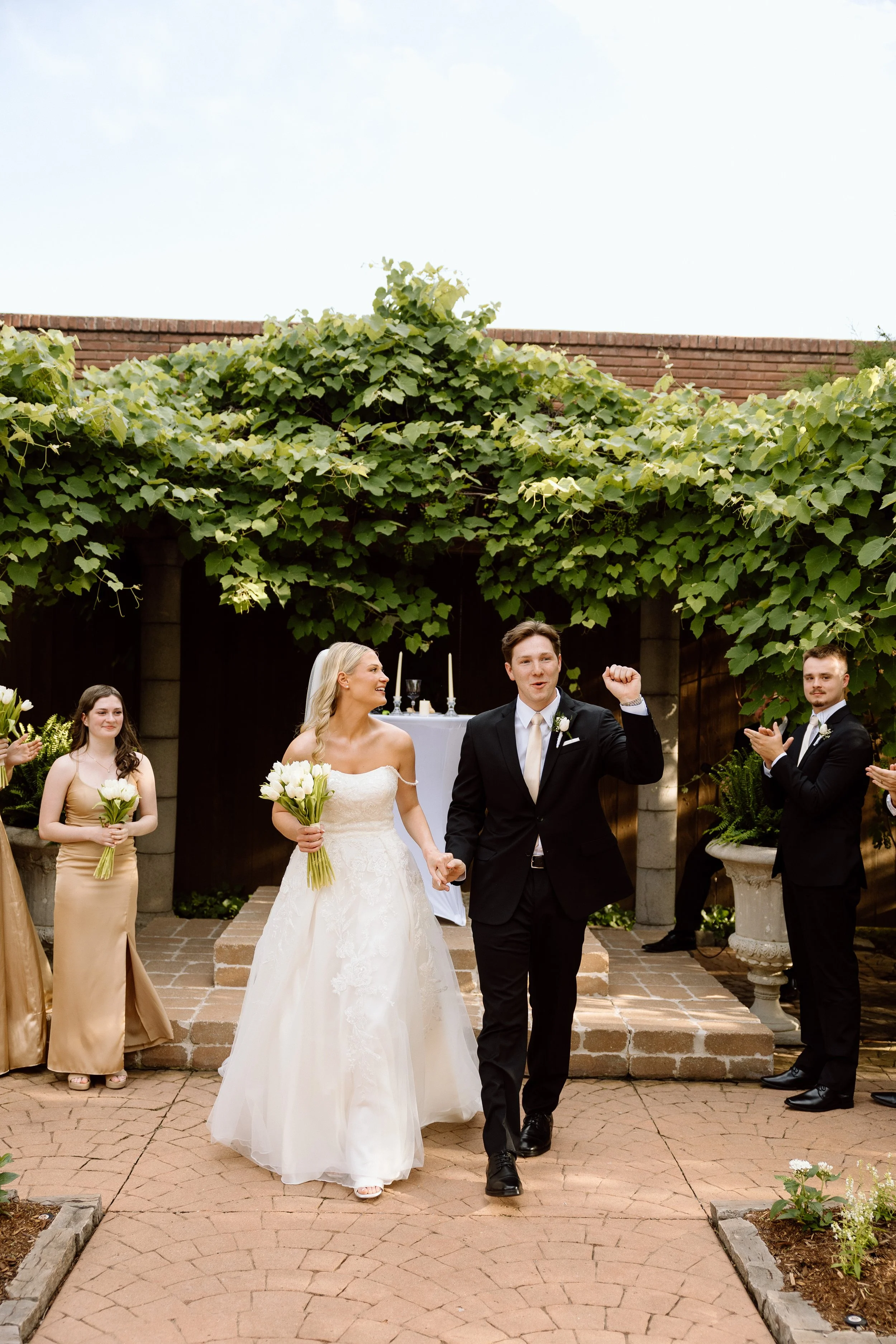 A wedding celebration outdoors with a bride in a white wedding dress holding a bouquet of white flowers, a groom in a black suit making a fist gesture, and several bridesmaids and groomsmen in formal attire clapping and smiling under a green leafy ca