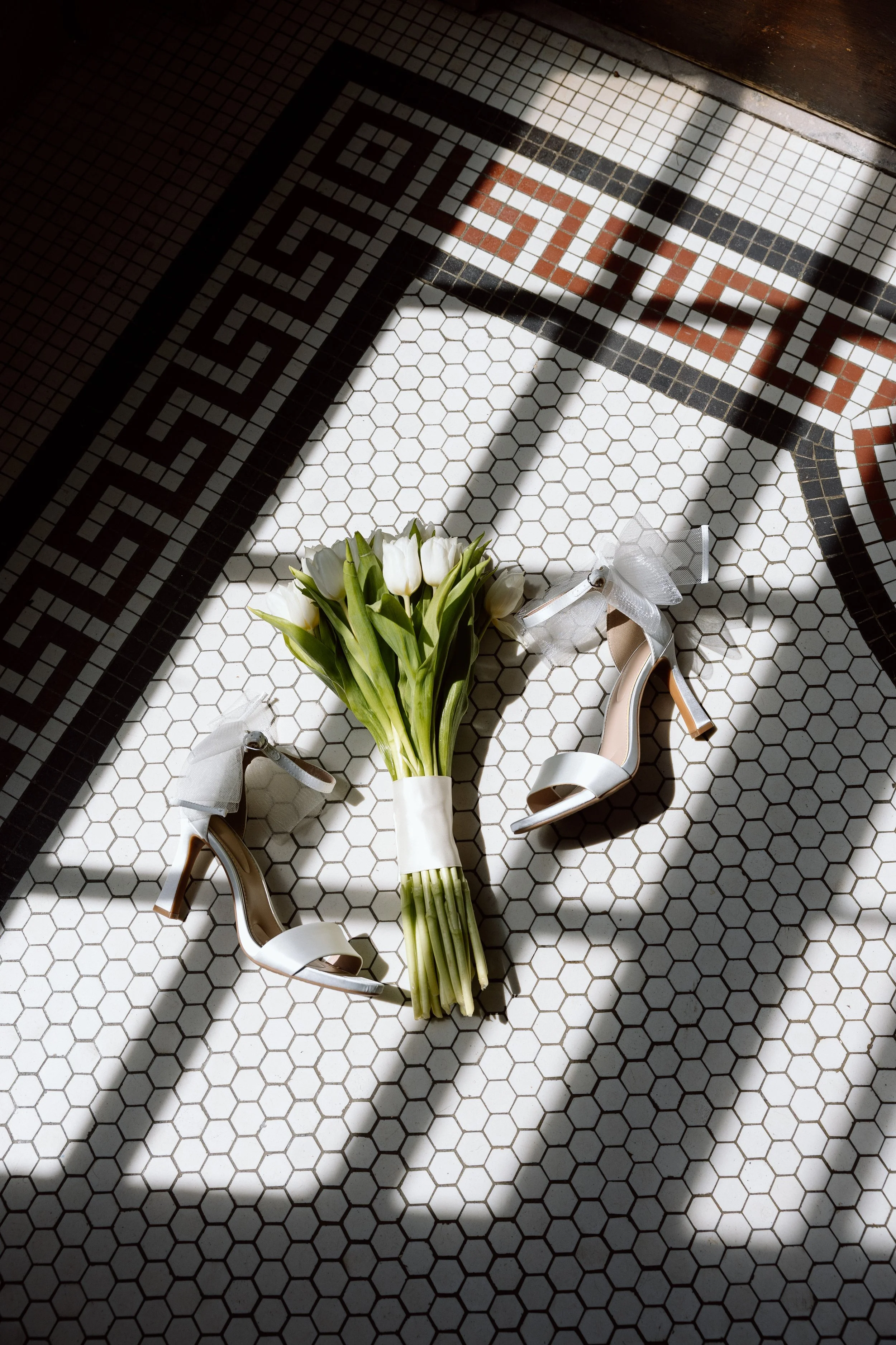 A bouquet of white tulips tied with a ribbon alongside a pair of white high heel shoes on a tiled floor with geometric black and white patterns.