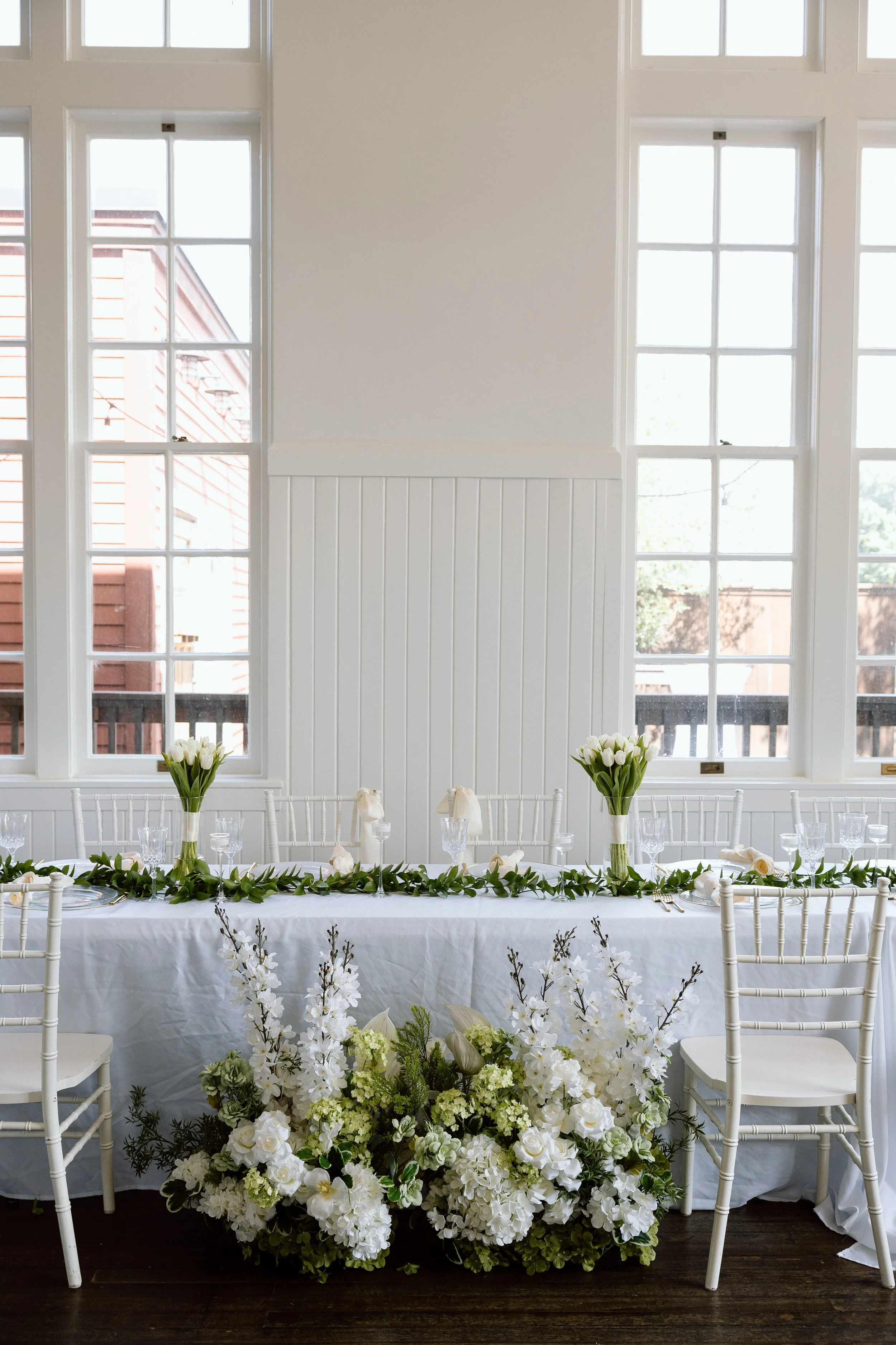 A long table set for a wedding reception with white chairs, floral centerpieces, and greenery on a white tablecloth. Behind the table are large windows with a view of neighboring buildings and a balcony outside.
