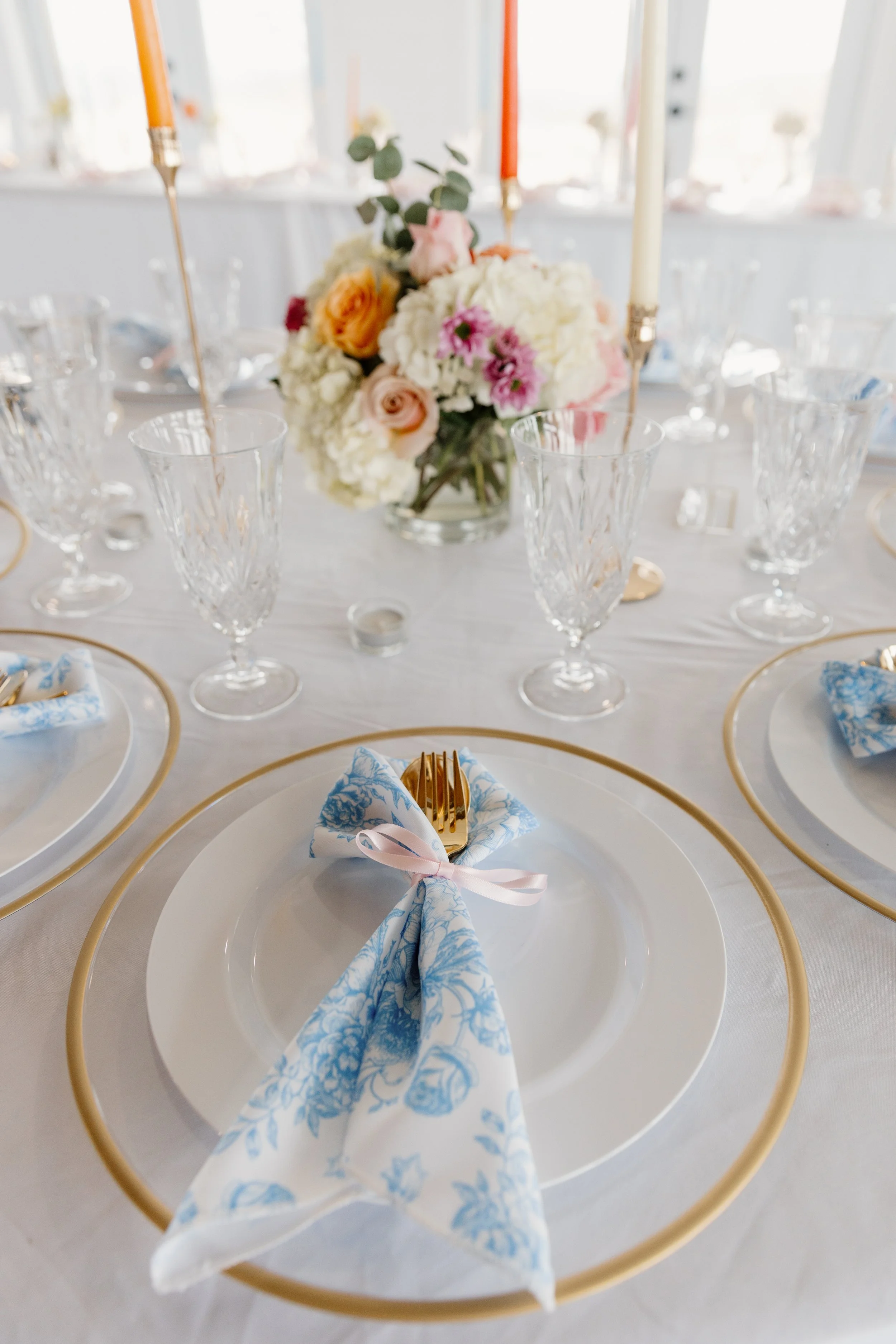 Table setting with a white plate, a blue patterned napkin tied with a pink ribbon, gold flatware, and a floral centerpiece of white, pink, orange, and purple flowers with green leaves. There are three tall candles in gold holders and crystal glasses 