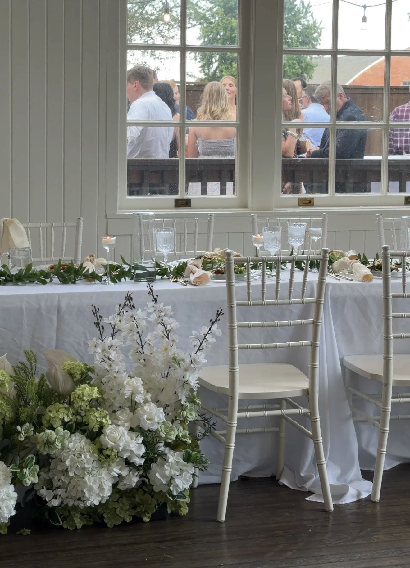 A decorated event table with white chairs and floral arrangements in front of a window overlooking a patio with people socializing outside.