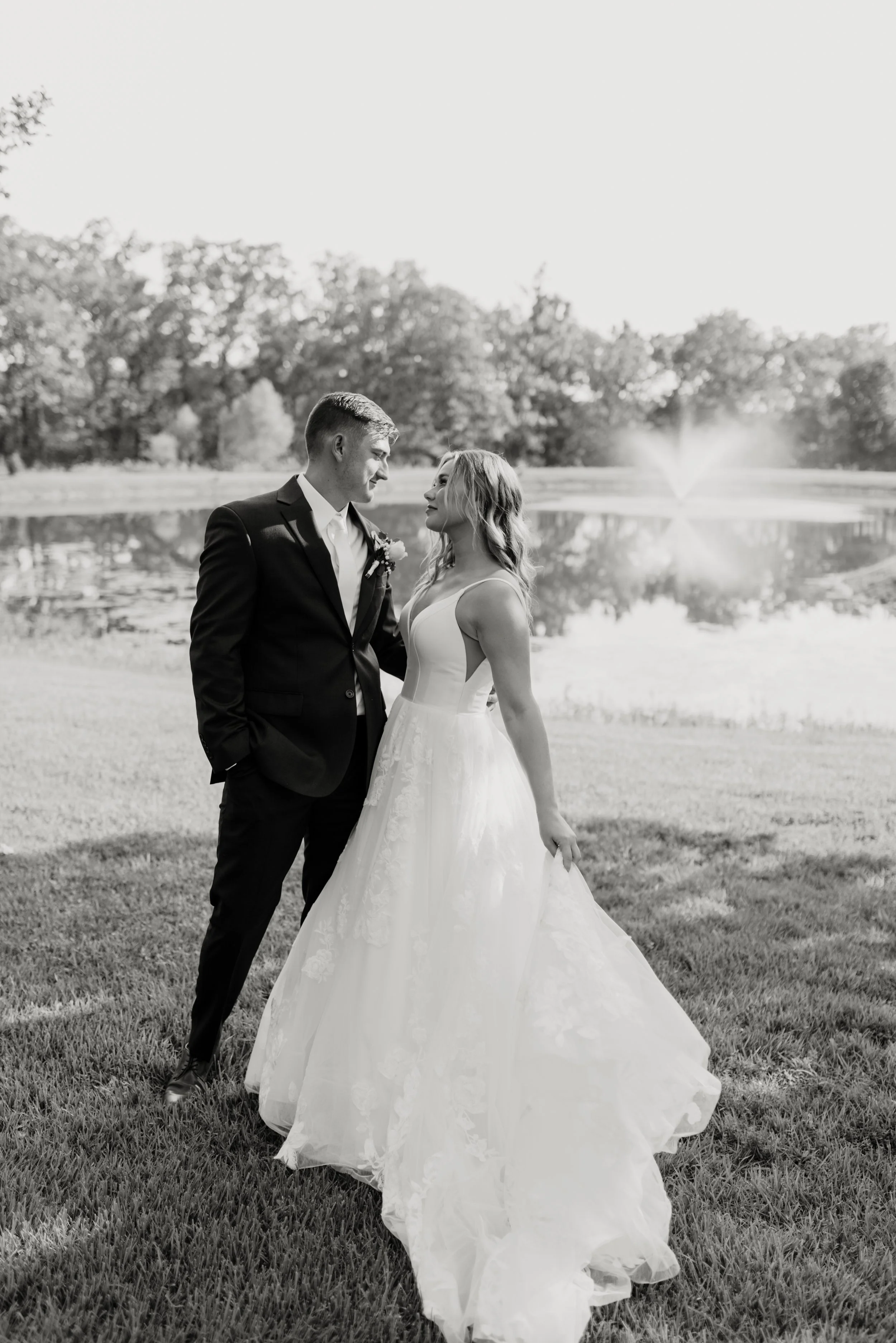 A black-and-white photo of a bride and groom standing by a pond with a fountain, trees in the background, and holding hands, dressed in wedding attire.