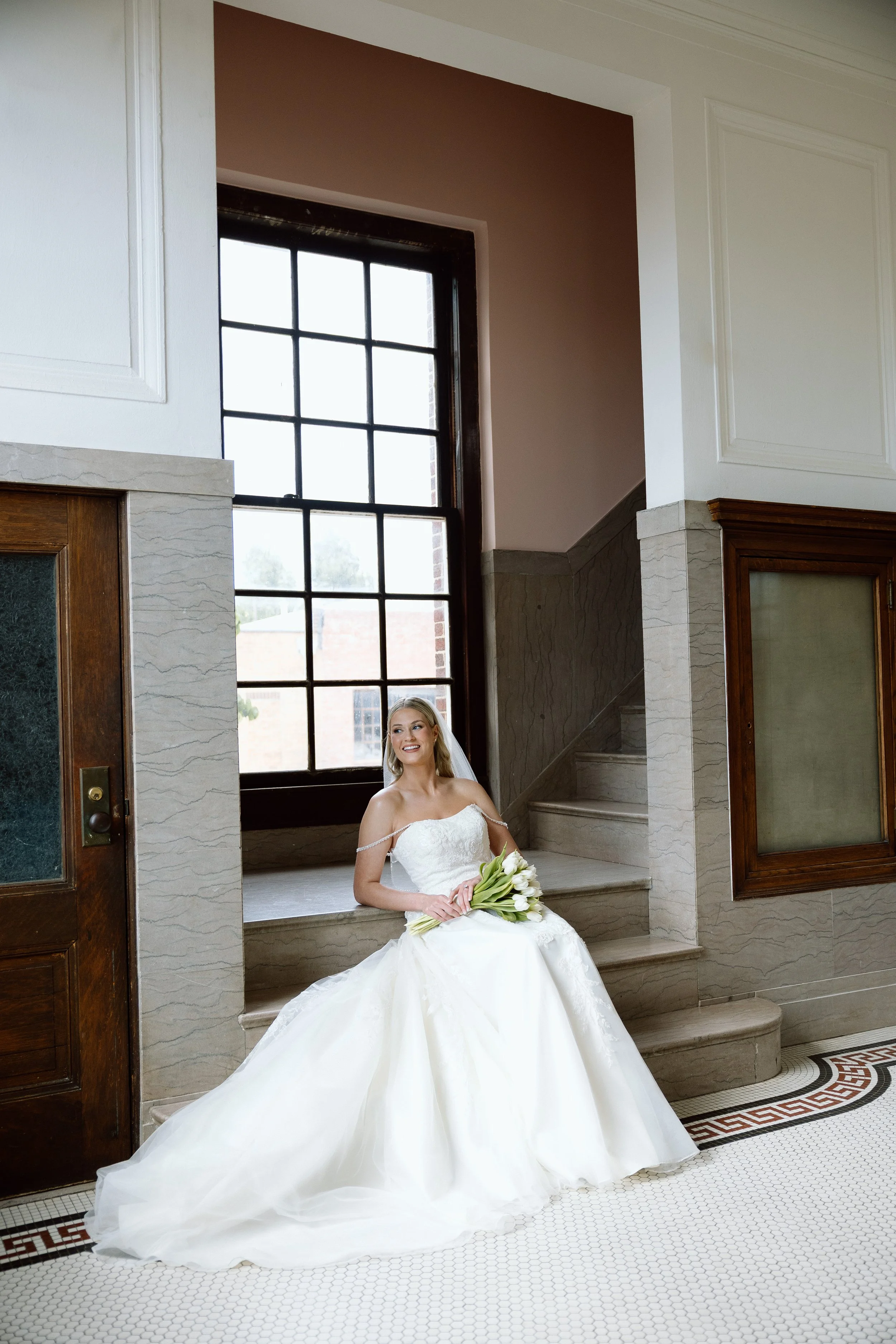 A smiling bride in a strapless white wedding gown sitting on stairs inside a building with large windows, holding a bouquet of white tulips.