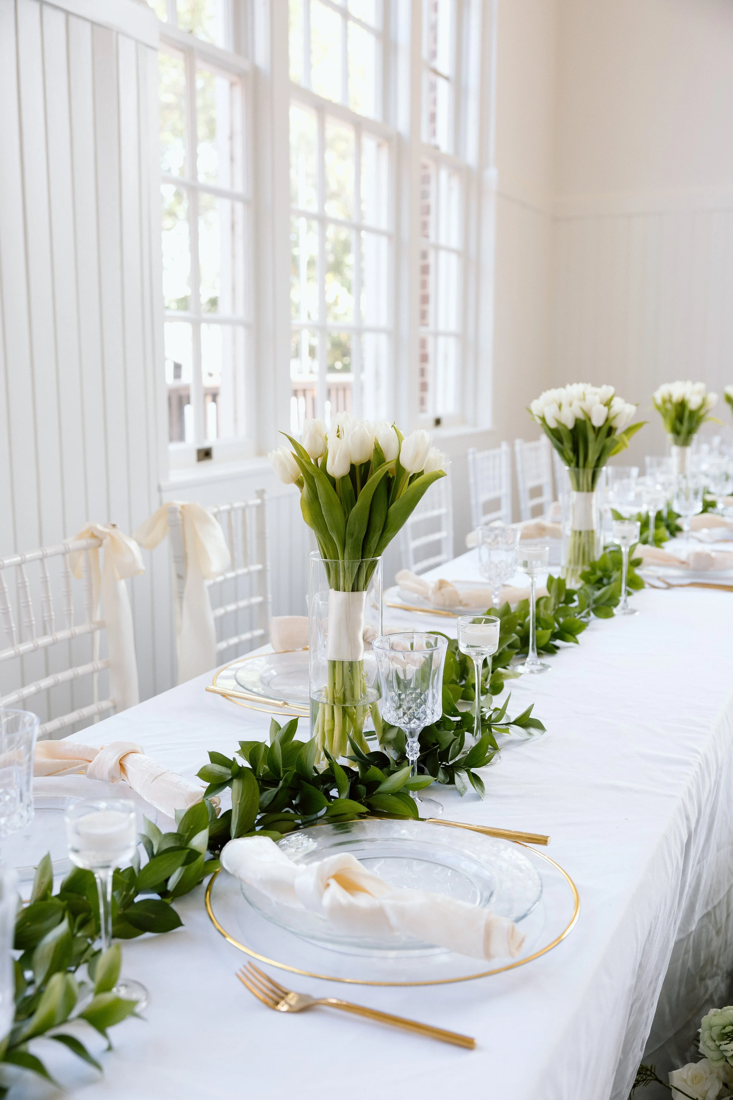 Elegant wedding reception table decorated with white and green floral arrangements, white napkins, gold cutlery, and glassware, set near large windows letting in natural light.