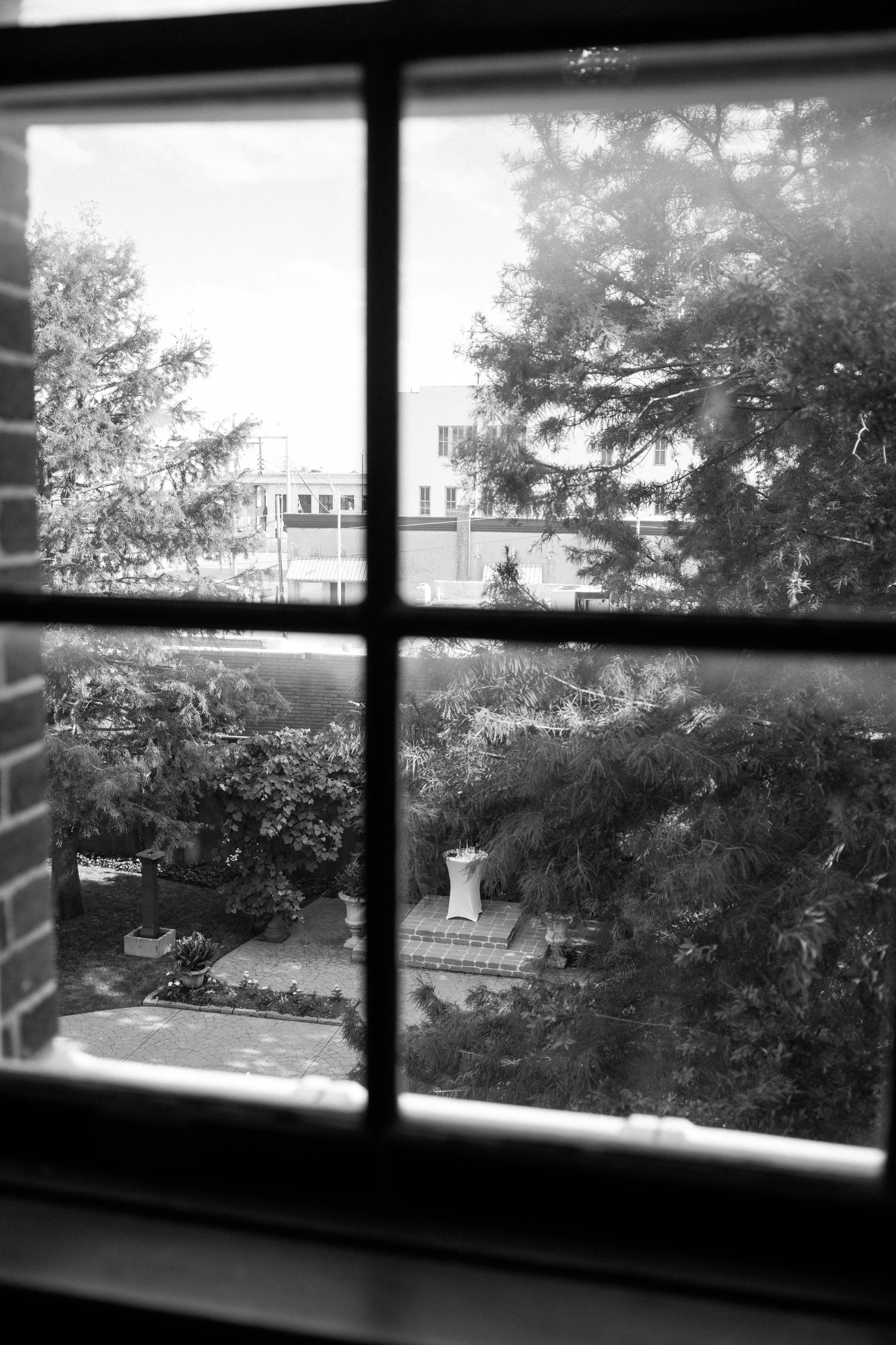 View through window blinds showing trees, a building, and a patio with potted plants and outdoor furniture.