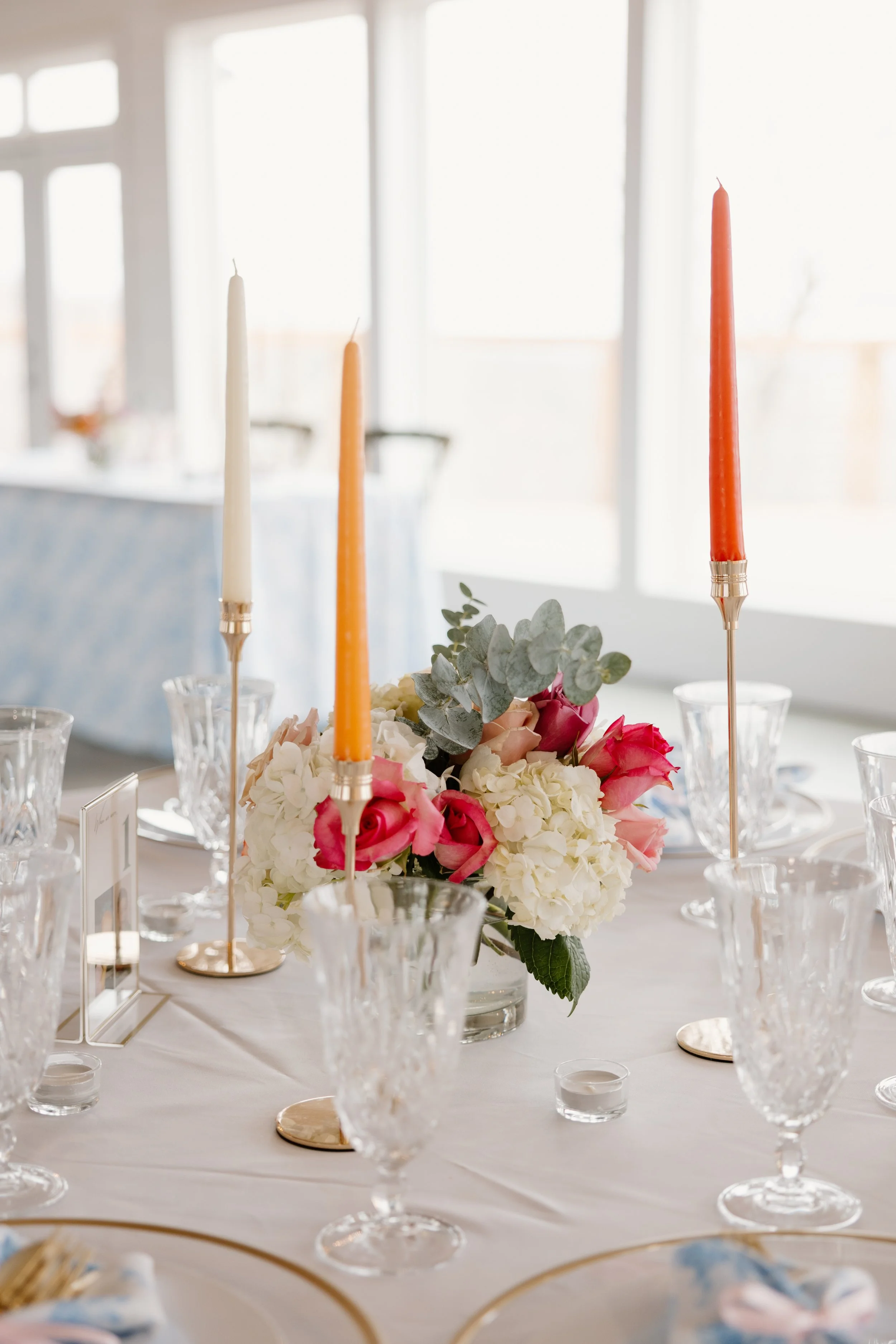 A floral centerpiece with pink and white flowers, greenery, and tall candles in orange and white on a table set for a celebration or wedding reception.