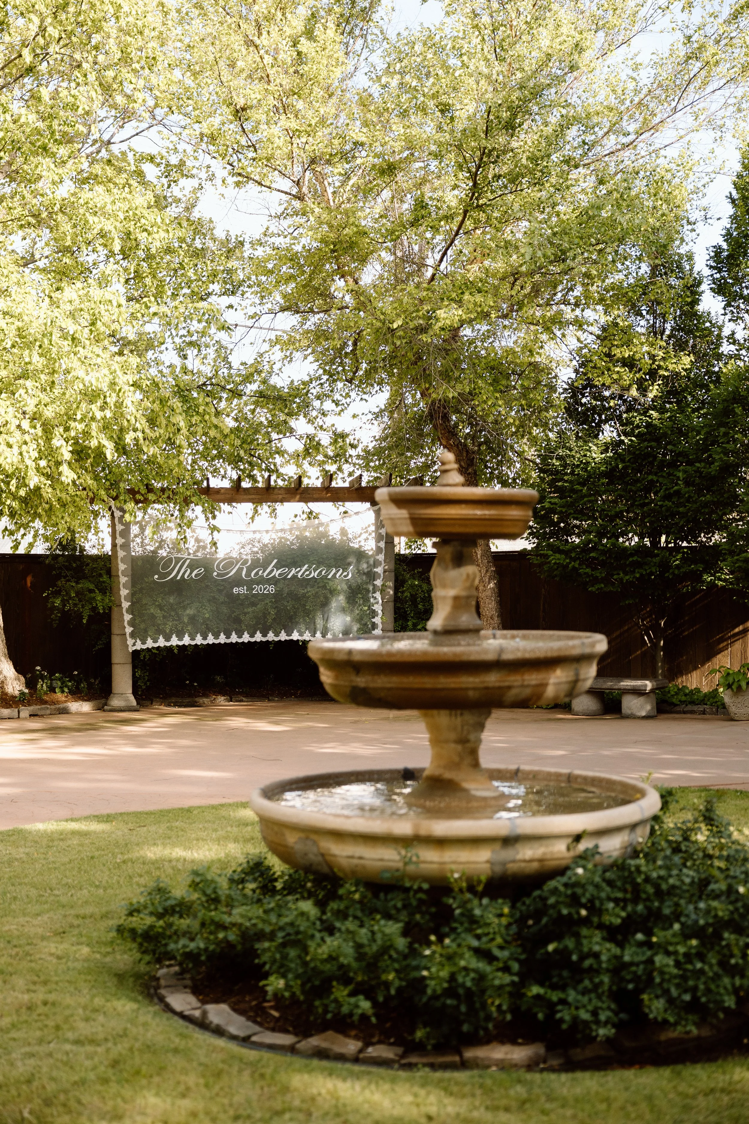 A two-tier fountain in the foreground, grass, bushes, and trees behind it, with a sign that reads "The Robertsons est. 2026" hanging on a fence in the background.