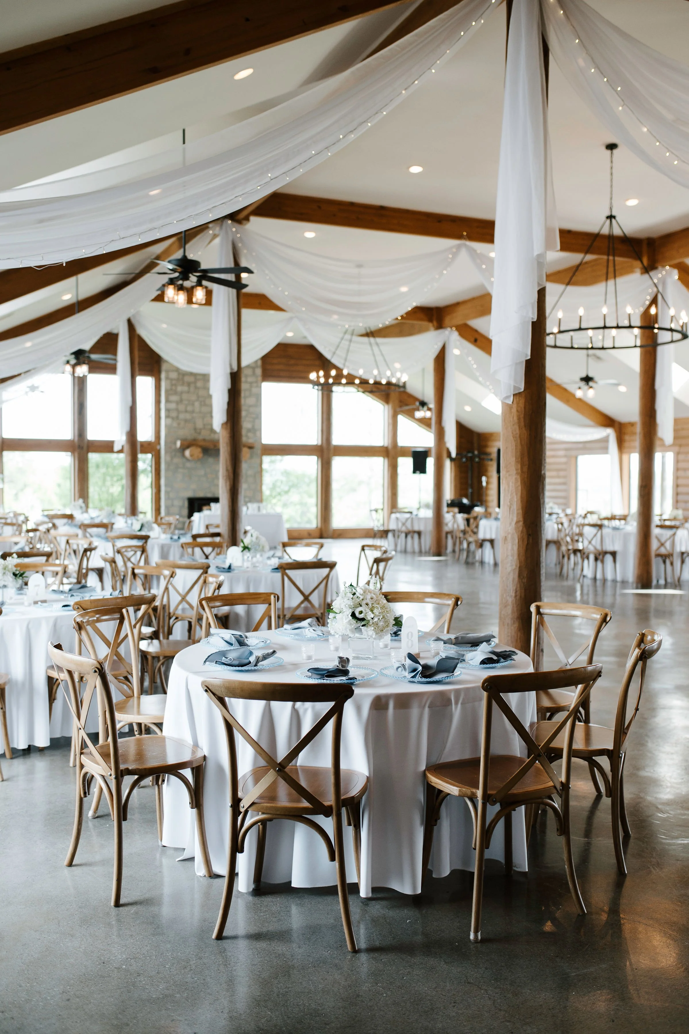 Elegant event hall decorated with white drapes and string lights hanging from wooden beams, set up for a formal occasion with round tables, white tablecloths, and flower centerpieces.