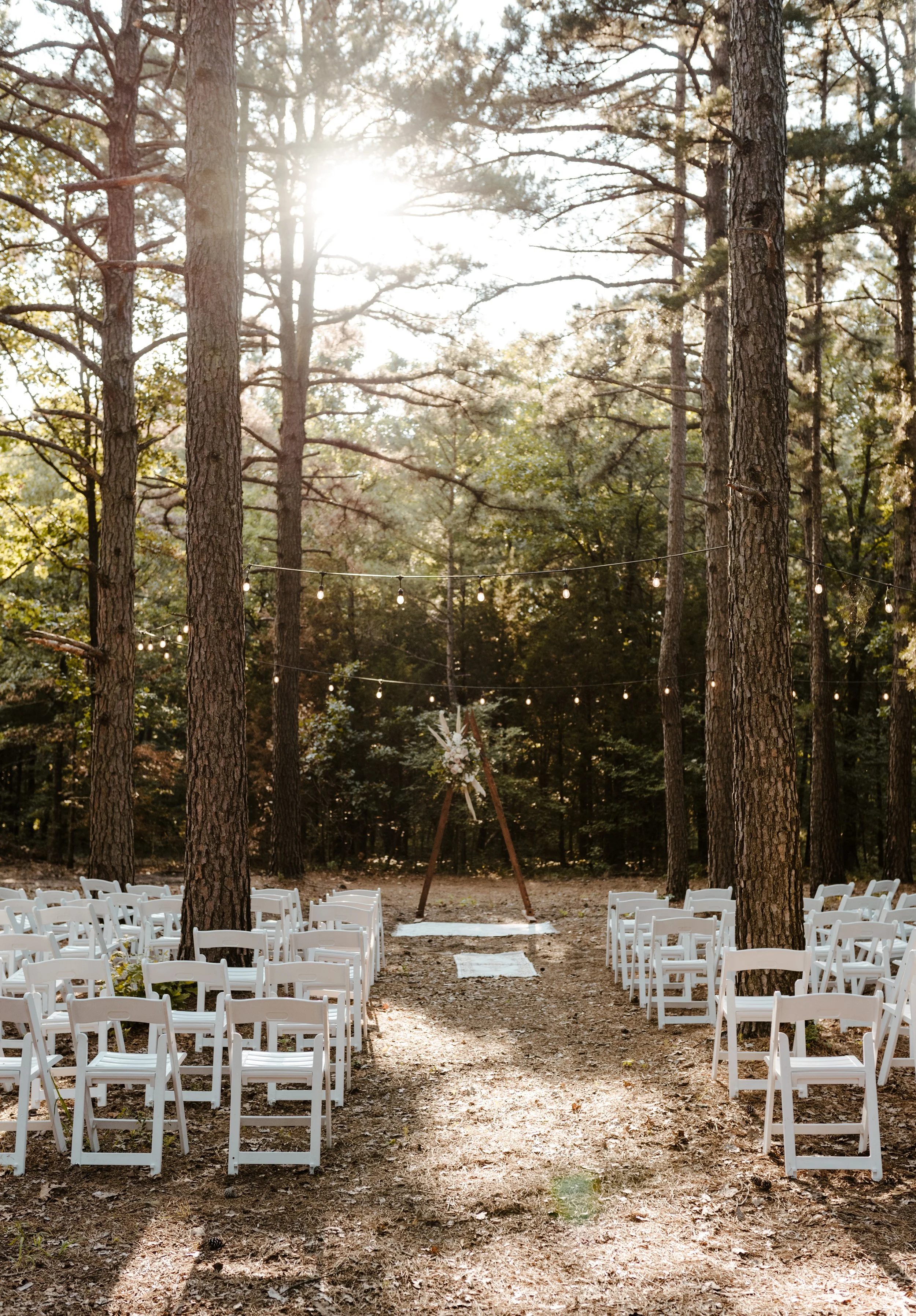 Outdoor wedding ceremony setup in a wooded area with white chairs arranged on either side of a dirt aisle, string lights overhead, and a floral arch at the altar, illuminated by sunlight filtering through trees.