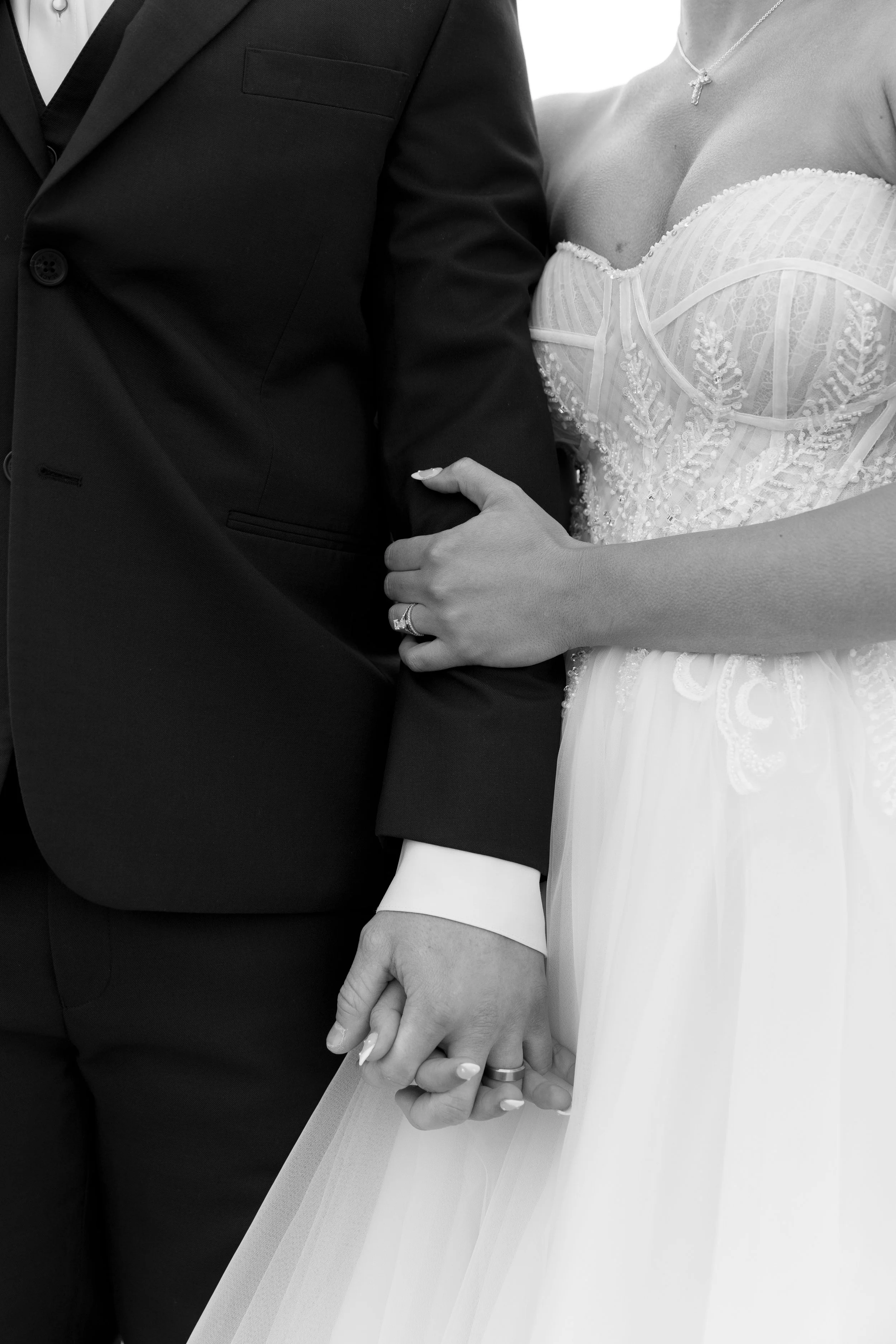Close-up of a bride and groom holding hands, focusing on the hands and part of their attire, with the bride wearing a wedding dress and the groom in a suit.