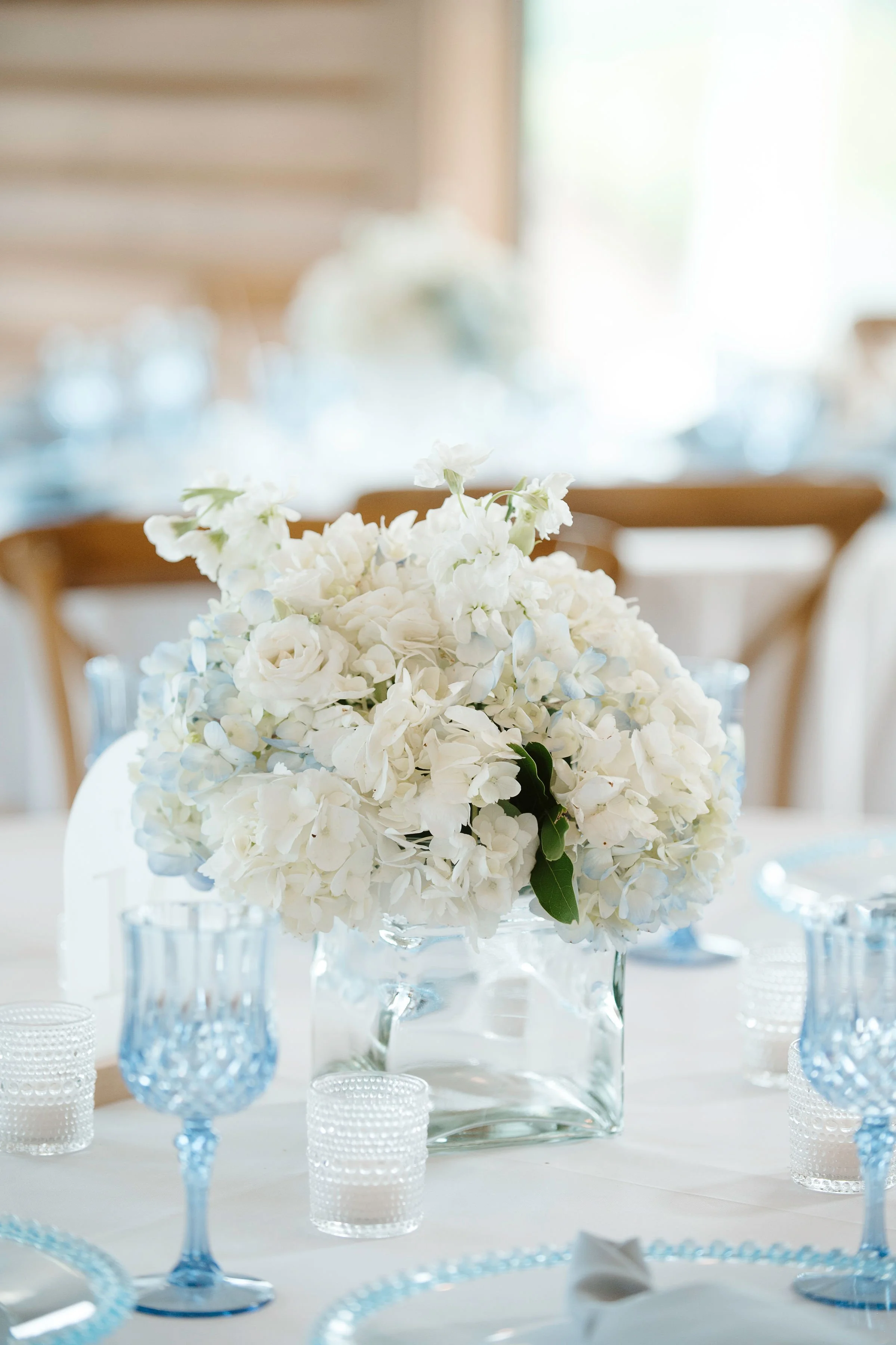 A floral centerpiece with white flowers in a glass vase on a table, surrounded by blue goblets and small candles, in a softly lit room.