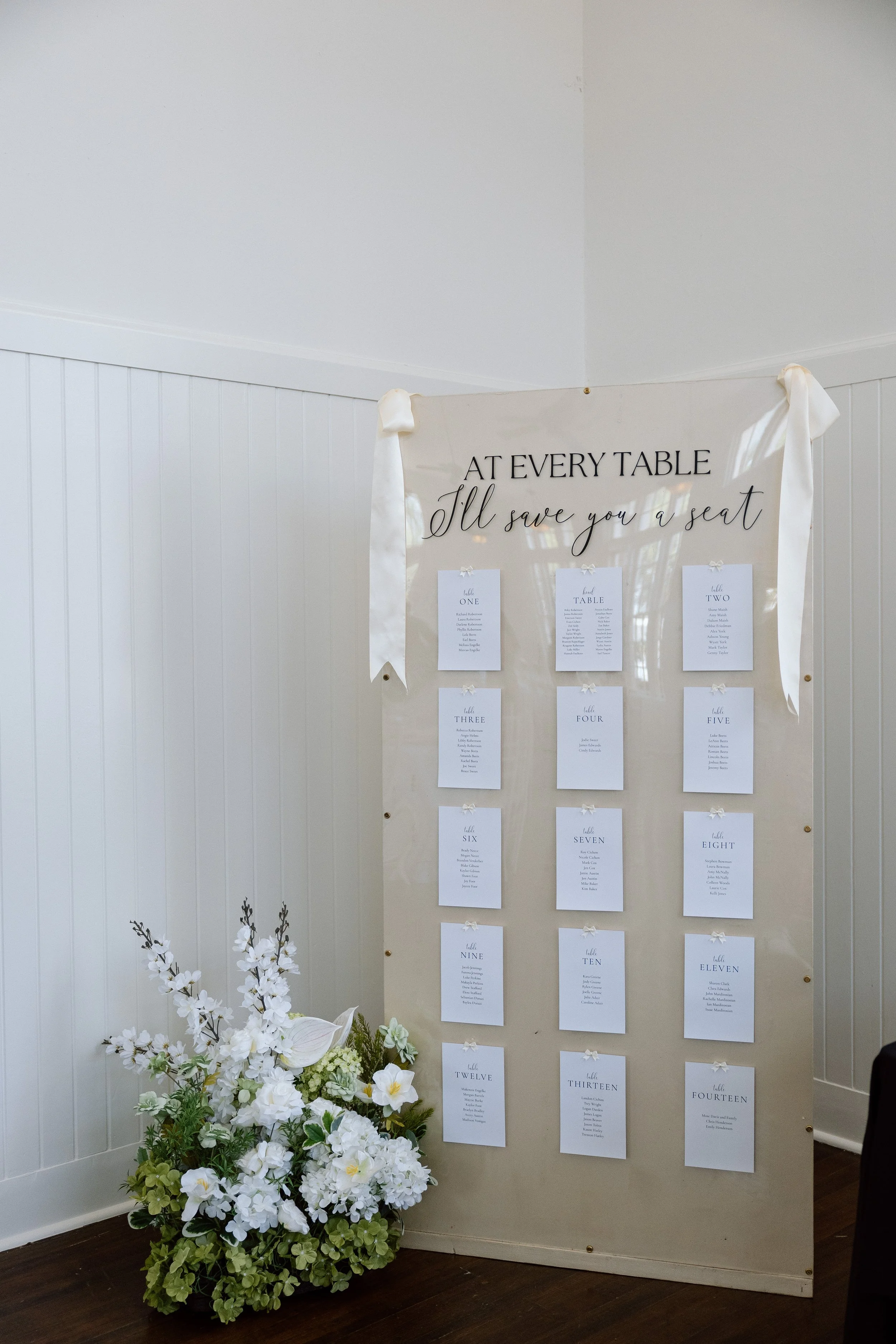 Seating chart for an event, with a floral arrangement in front, displaying tables labeled one through fourteen, each with a list of names.