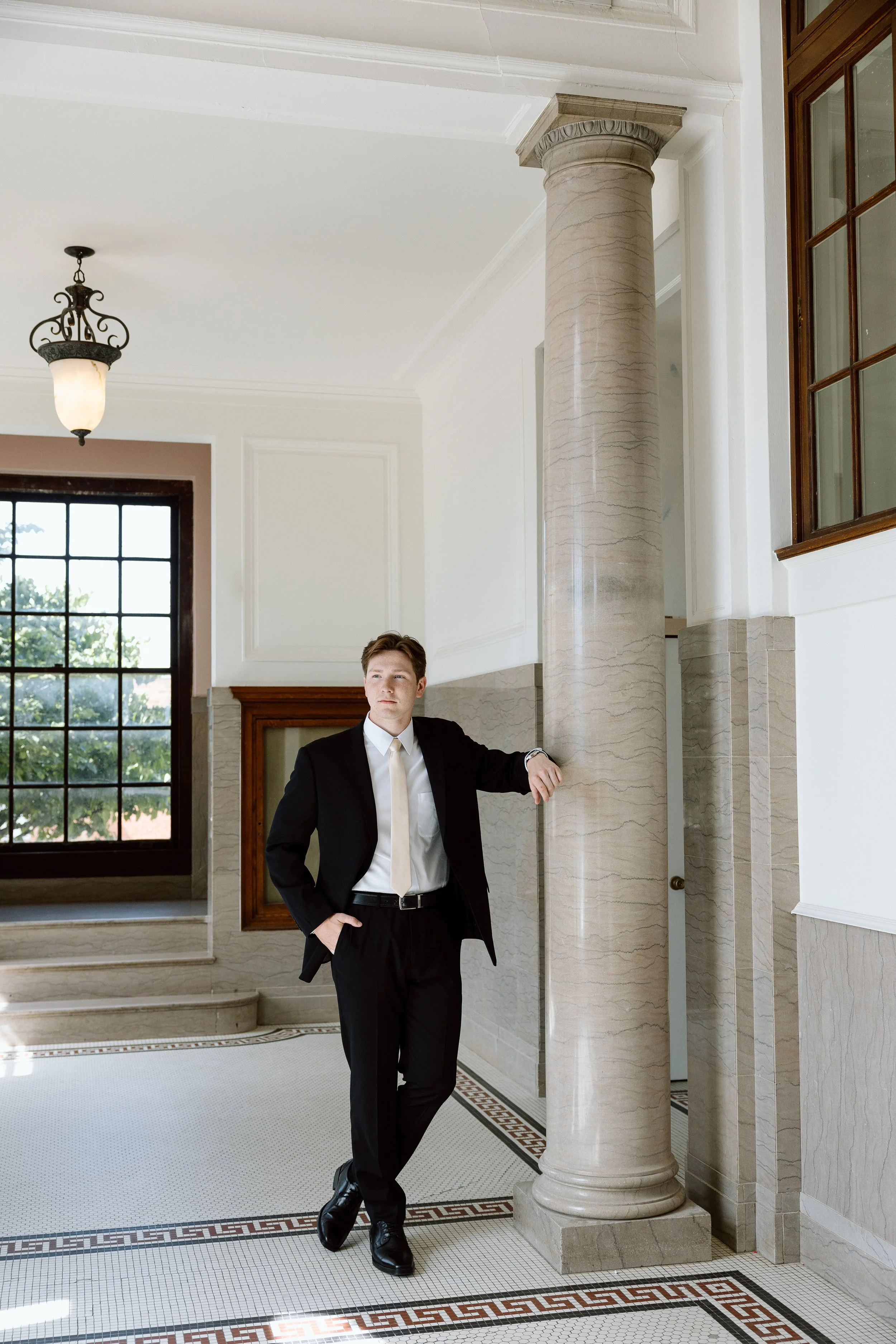 A young man in a black suit, white shirt, and beige tie standing in a luxurious, bright interior space. He rests one arm on a large marble column and the other hand in his pocket, looking thoughtfully to the side. The room features large windows, orn