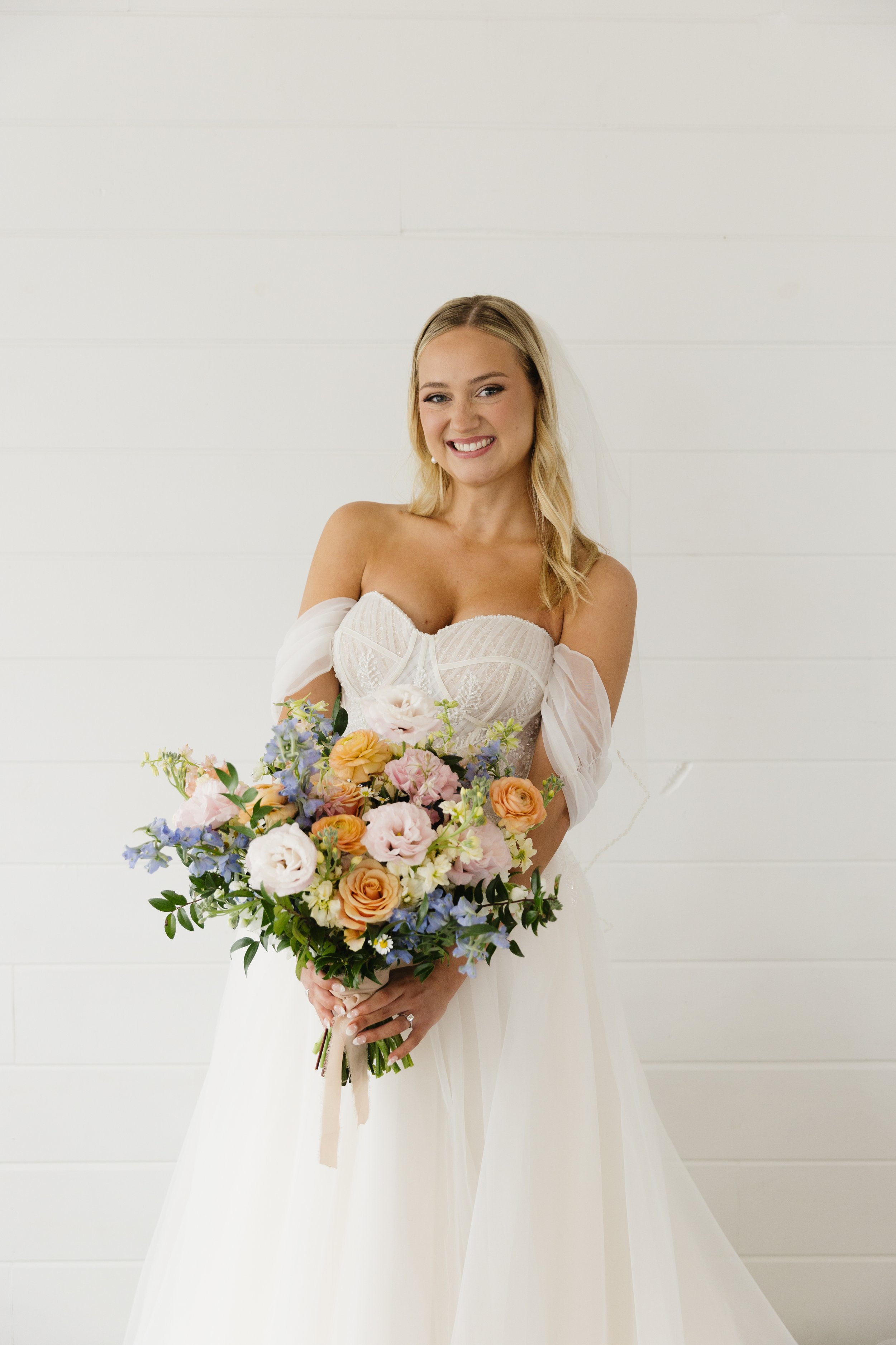 A young woman in a wedding dress holding a large bouquet of colorful flowers, smiling, standing against a white wall.